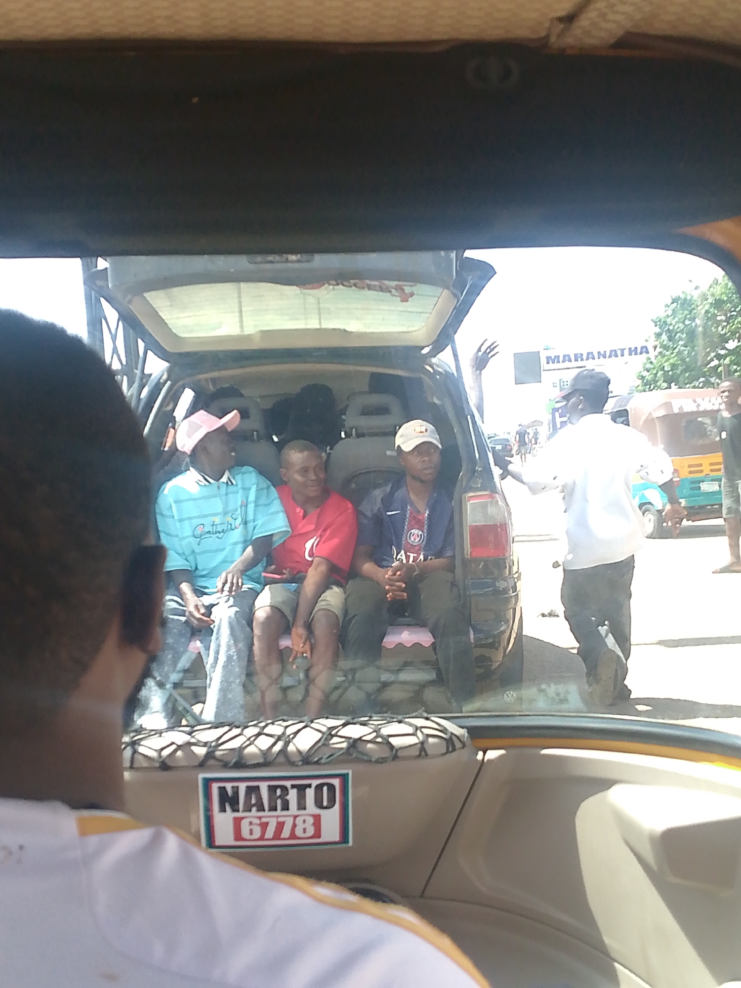In the city of Bukuru, Nigeria, four men are seated in the back of an open-trunk vehicle. One man, wearing a pink cap and a light blue horizontally striped shirt, is on the left. Next to him, a man in a red t-shirt is seated. Another man in a blue and white striped shirt and a dark baseball cap sits to the right. A fourth man, partially obscured, is seated behind the man in the red shirt. A sticker with the text "NARTO 6778" is visible on the back of the vehicle. A man in a white shirt is standing outside the vehicle, facing away from the camera, with his arm raised. In the background, a building with the sign "MARANATHA" is visible. A three-wheeled auto-rickshaw is parked to the right of the building. The scene appears to be on a paved street under a bright sky.