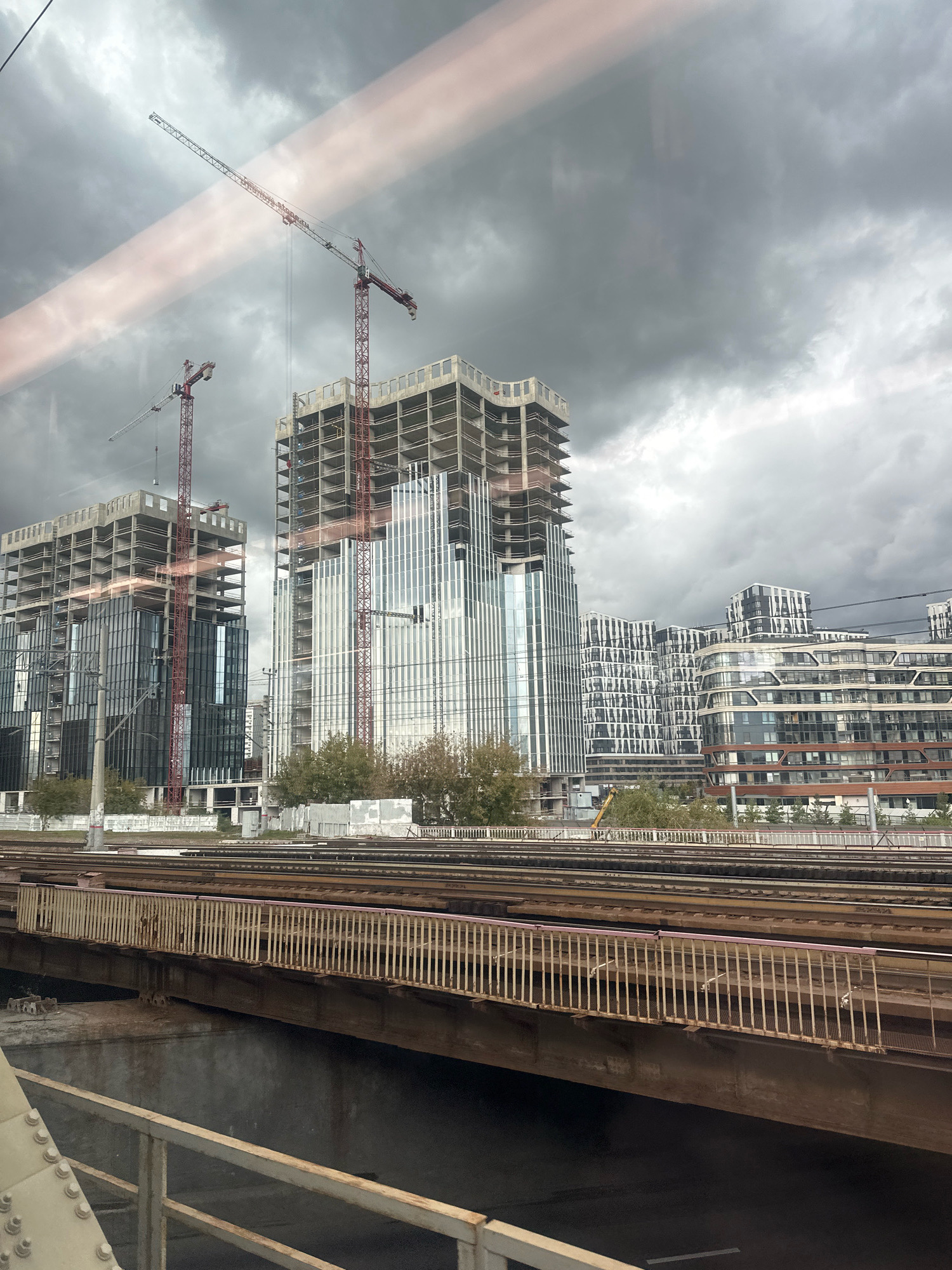 The image shows a construction site with several high-rise buildings under construction, as well as completed buildings in the background. Two large red construction cranes are visible, one towering over the tallest unfinished building and another smaller one to its left. The buildings are a mix of glass facades and concrete structures. In the foreground, there are railway tracks and a bridge, suggesting the photo was taken from a moving train or another elevated vantage point. The sky is overcast with dark, cloudy conditions. The architectural style of the completed buildings in the background appears modern and varied, with some featuring curved facades and a pattern of windows. The scene is indicative of urban development and infrastructure. This scene is located in Moscow, Russia.