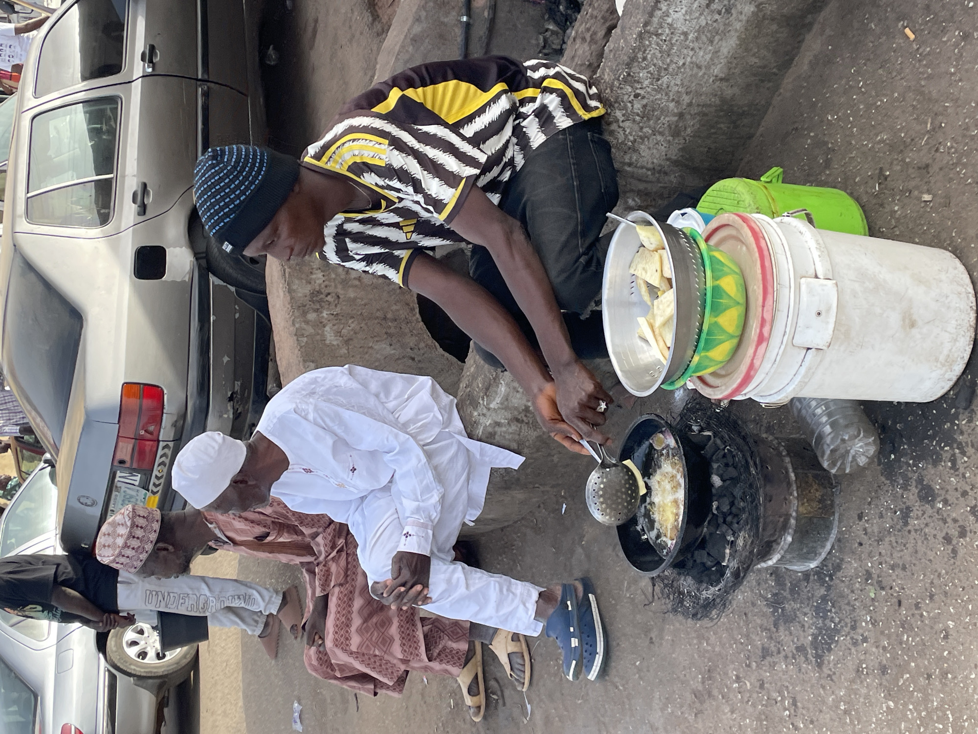 This image, captured in Jos, Nigeria, depicts an outdoor cooking scene where a street food vendor prepares food on a charcoal stove while several men observe or wait.
In the foreground, a man, dressed in a black beanie with blue stripes, a black, yellow, and white striped shirt, and dark trousers, is focused on frying food. He kneels or squats beside a black pan, which rests on a makeshift charcoal stove fueled by coals. He uses a slotted metal spoon to stir pieces of food, likely yam or potatoes, sizzling in hot oil.
Adjacent to the cooking setup, on his right, are various containers: a white plastic colander holding raw food chunks, a green bowl, and a large white bucket. A clear plastic bottle is also visible on the ground nearby.
To the left of the vendor, two other men are seated on the ground. One is dressed in a white traditional robe and a white cap, while the other wears a brown patterned traditional outfit and a matching cap, paired with light-colored pants that have the word "UNDERGROUND" printed prominently on one leg. Their footwear includes sandals and blue Crocs-style shoes.
In the background, an older model silver car is parked, with its rear bumper and a partially visible "LAGOS" license plate discernible. The ground beneath them is a rough, unpaved surface, possibly dirt or worn asphalt. The scene portrays a casual, outdoor food preparation and consumption setting, likely on a street or in a market area during daylight hours.