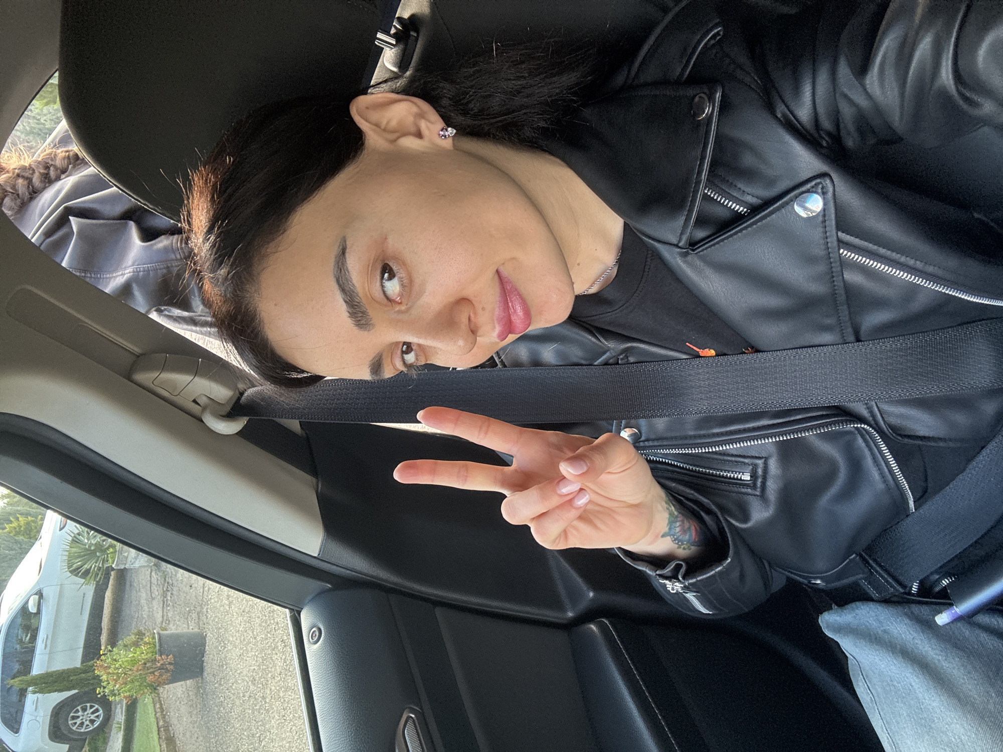 A person is inside a vehicle, wearing a black leather jacket and jeans. The person is making a peace sign with their right hand. The person has dark hair and a silver earring on their left ear. The person is wearing a seatbelt. A white car is visible outside the vehicle, along with a potted plant. The location is likely not Tbilisi, Georgia, as there is no discernible evidence to suggest this.