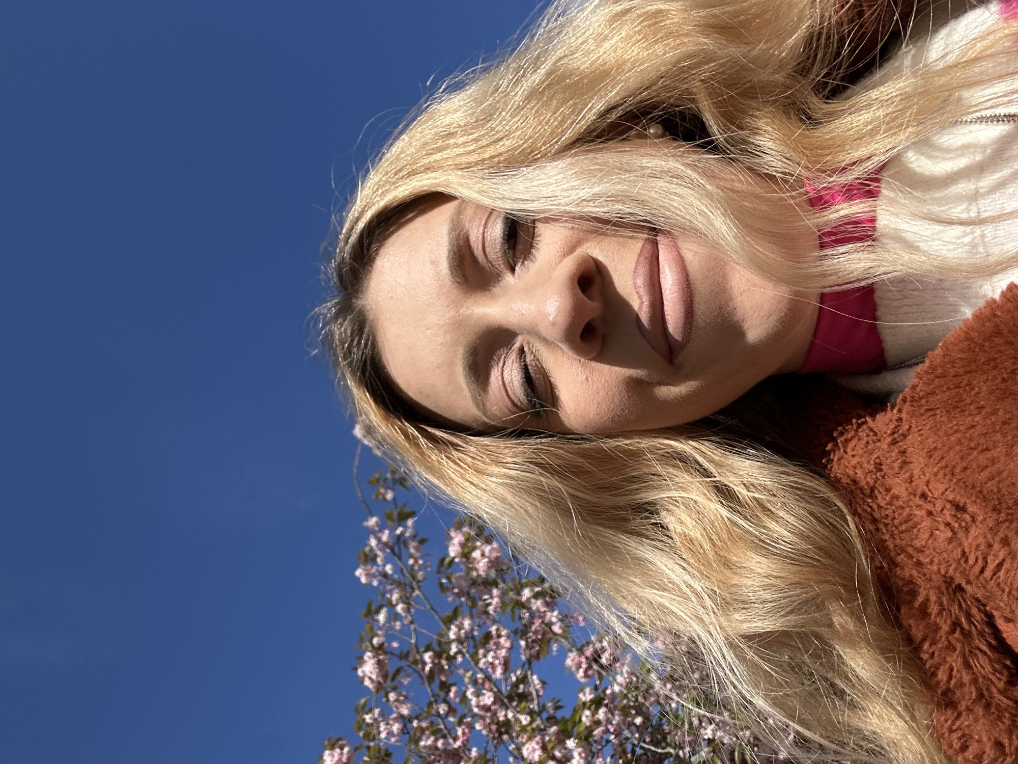 A low-angle, close-up selfie captures a young woman with long, wavy blonde hair, her face angled upwards. Her eyes are gently closed or cast downwards, and a subtle smile plays on her lips, conveying a sense of peace and contentment. She wears light makeup, including eyeshadow and neutral-toned lipstick. Partially visible is a white and pink striped garment beneath a fluffy, reddish-brown coat or jacket. The background features a bright, clear blue sky, with a blossoming tree covered in small pink flowers visible on the left side of the frame. The scene is bathed in bright daylight, indicating a sunny day, likely in spring given the blossoms. While not explicitly shown in the image, the context suggests this scene is in Amsterdam, Netherlands. No text is visible in the image.