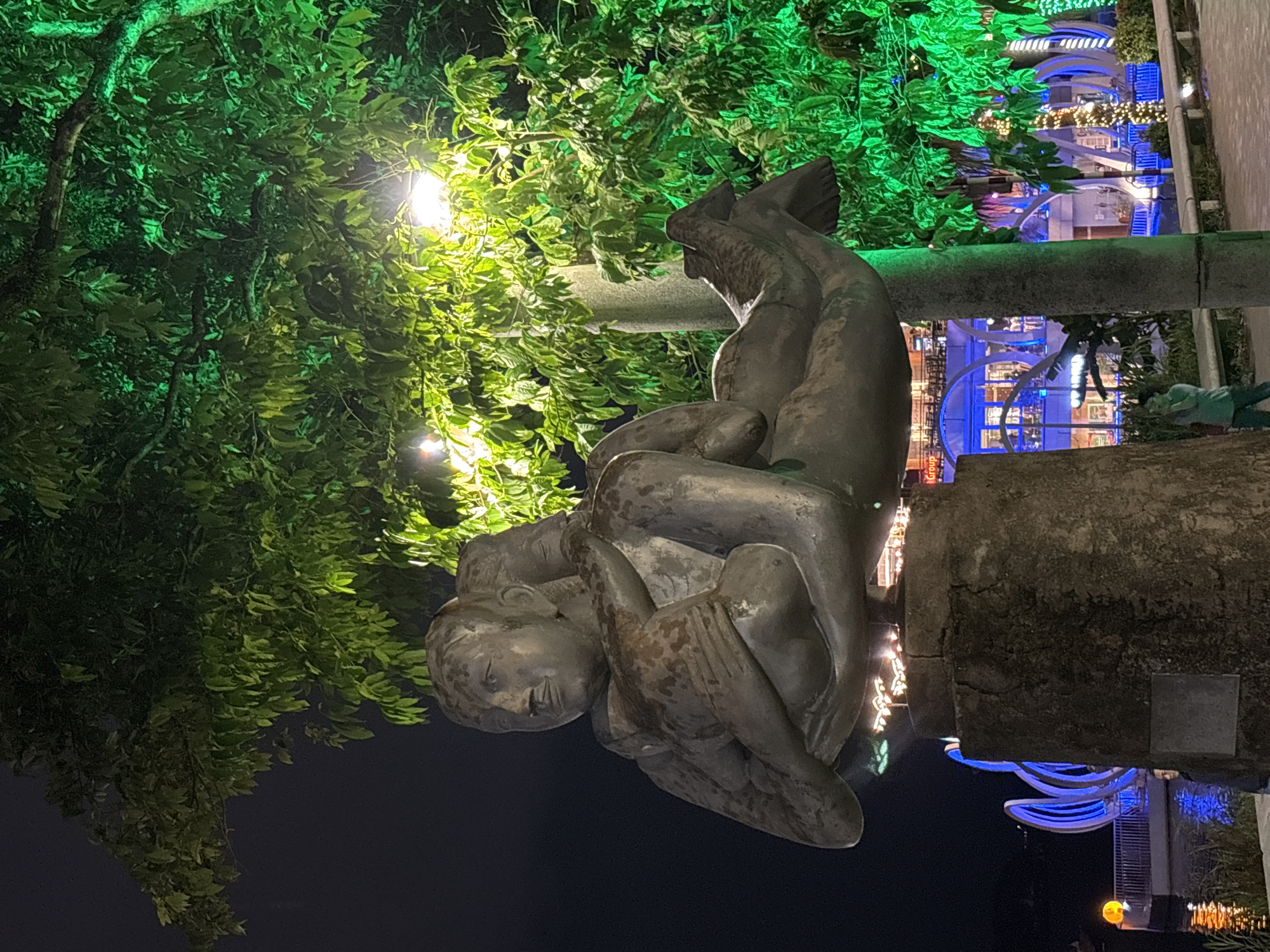 The image depicts a nighttime scene in Thuận Hóa Ward, Vietnam, featuring a bronze sculpture of two figures embracing, positioned on a concrete pedestal. The sculpture is illuminated by green and yellow lights, which also highlight the lush foliage of trees surrounding it. In the background, a body of water is visible with illuminated structures and decorative lights, suggesting a waterfront area. A person, dressed in a teal shirt and dark pants, is standing near the sculpture, looking at their phone. The overall atmosphere is festive and illuminated, characteristic of a public space at night.