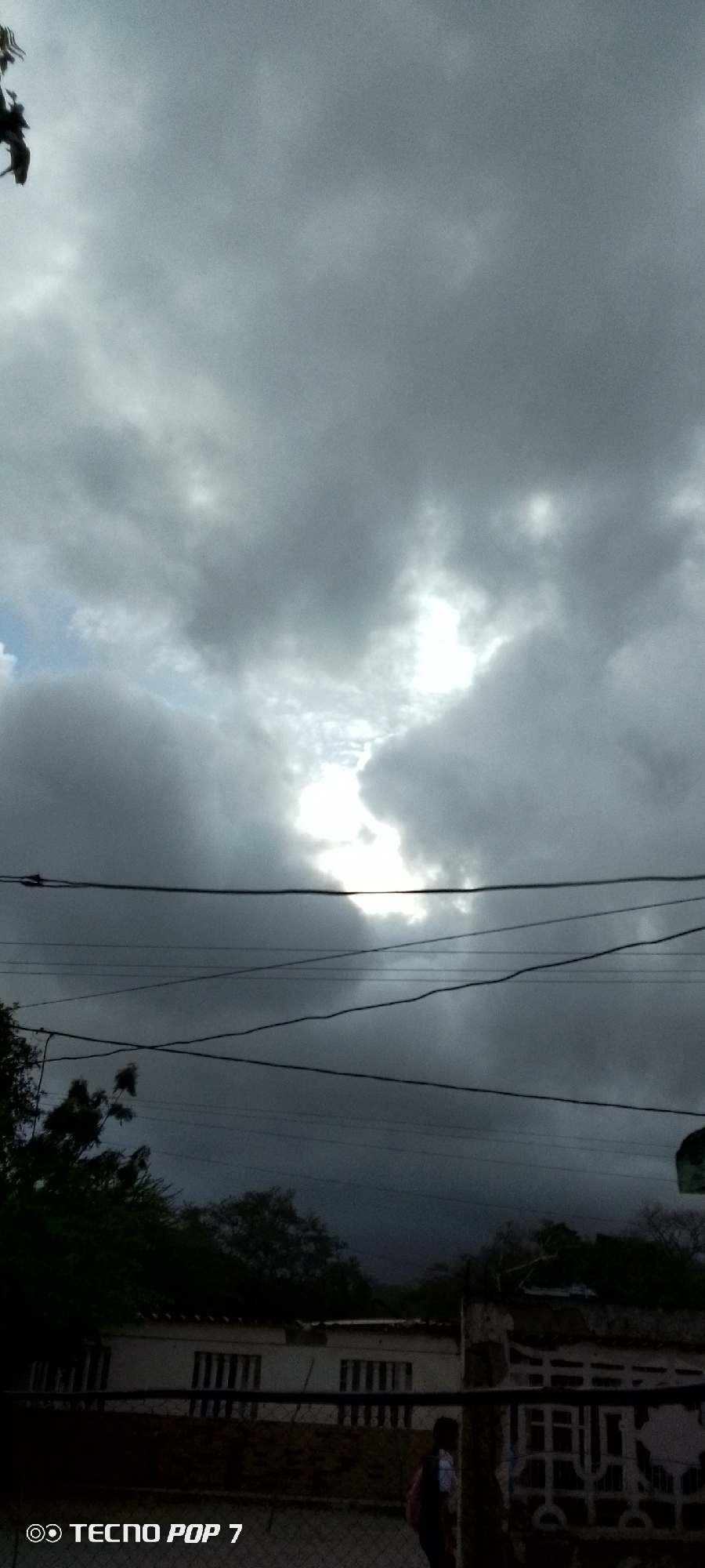 This is a vertical, outdoor, low-angle shot of a cloudy sky over a residential area. In the foreground, several dark power lines crisscross the frame horizontally. Below the power lines, there is a glimpse of trees and parts of buildings, including a white structure with windows and a fence. The sky is filled with heavy, grey clouds, with a bright patch of sunlight breaking through in the upper center. The lighting suggests it may be late afternoon or early evening, with an overcast or stormy weather condition. A person, partially visible from behind with a backpack, is walking on the right side of the frame near a building. The overall mood is somber due to the dark clouds. The bottom left corner of the image contains the text "© TECNO POP 7". The location context provided is Altagracia, Venezuela.