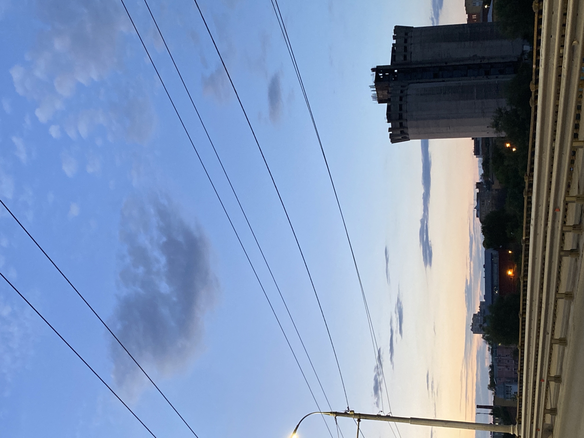 This media file is a photograph of a cityscape at dusk. The image is oriented vertically, with the sky occupying the majority of the frame. Several power lines are visible, stretching diagonally across the sky. In the background, a large, industrial-looking concrete structure, possibly a silo or a disused factory, dominates the right side of the frame. Below this structure, the silhouettes of trees and buildings are visible, with a warm light emanating from one of the buildings, suggesting it is illuminated from within. The sky is a gradient of blues and oranges, typical of a sunset or twilight. Scattered clouds are present in the sky, some appearing as dark smudges and others as lighter wisps. The overall mood of the image is somewhat somber and industrial, with a touch of urban decay implied by the appearance of the large concrete building. The photograph appears to have been taken from an elevated position, possibly a bridge, as indicated by the railing on the right edge of the image. There is no visible text in the image.