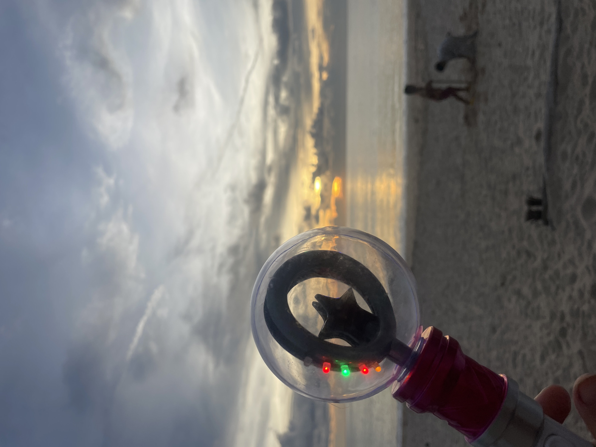 The image shows a beach scene at sunset, possibly in Choeng Thale, Thailand. In the foreground, a person holds a plastic bubble wand with a star-shaped center and LED lights (red, green, and orange). The background features a sandy beach, the ocean, and a cloudy sky with the setting sun. Figures of two people and a dog are visible in the distance on the beach.