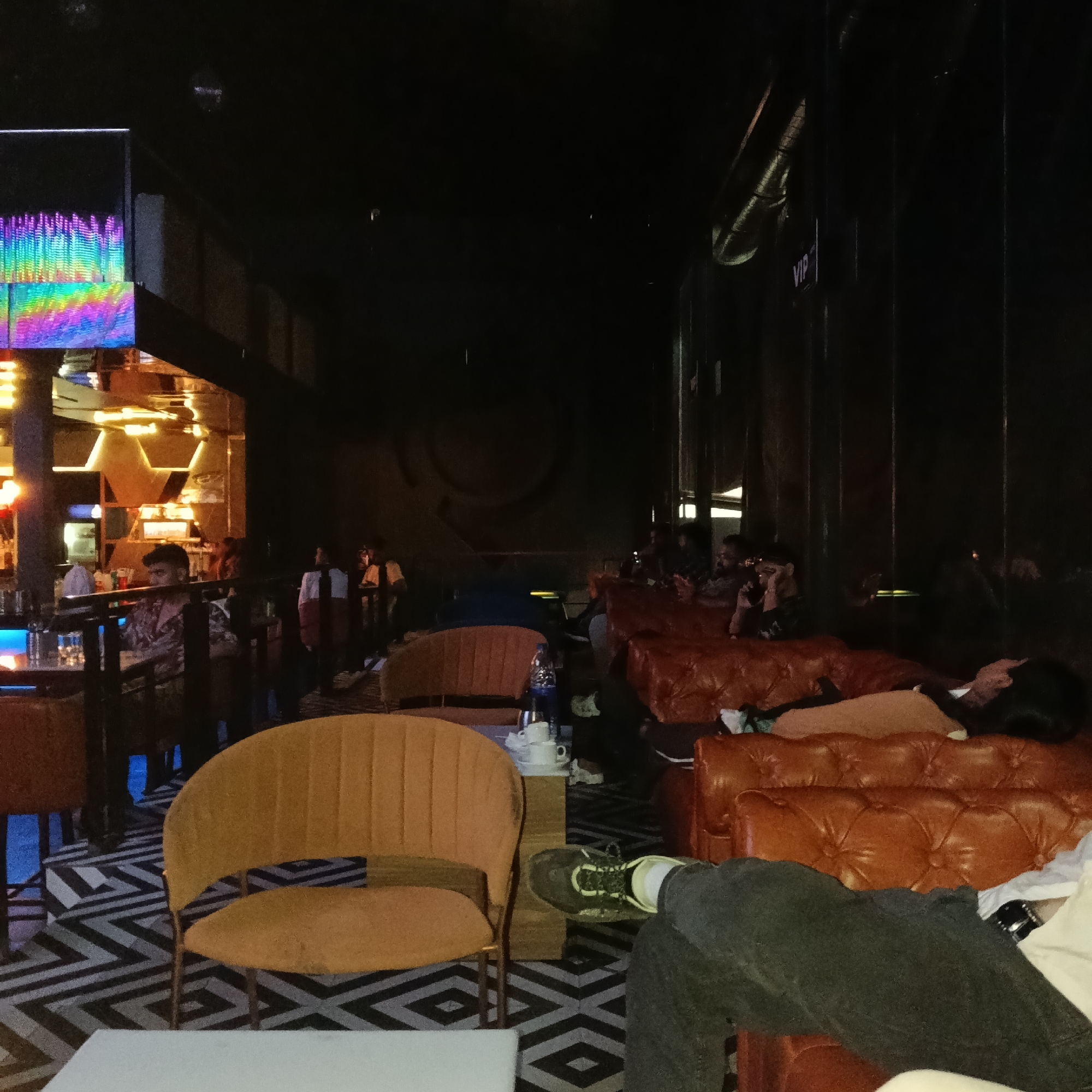 The scene appears to be inside a lounge or bar, likely in Sinquerim, India, based on the user's prompt. The lounge is dimly lit. There is a bar area visible on the left side of the image, with bartenders and patrons. A screen displays a colorful equalizer graphic. Further into the room, there are several orange leather couches and chairs where people are relaxing. Some people are sitting up, while others are reclining or lying down. There is a table with cups and a bottle of water. The floor has a black and white geometric pattern. Some areas have "VIP" written on the walls. There are people on the couches.