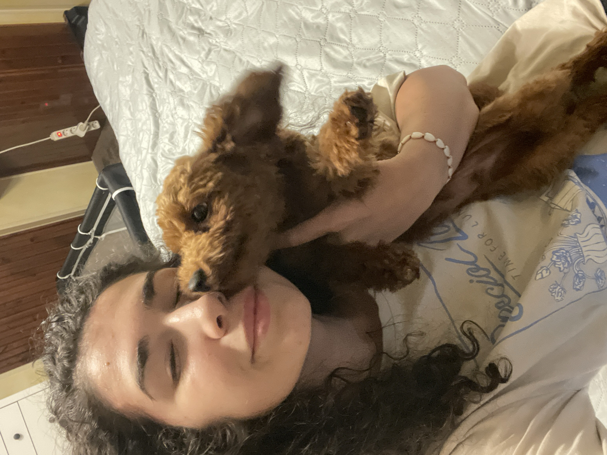 A woman is lying on a bed with a light gray, quilted comforter. She is on her back, with her eyes closed and a slight smile. Her curly dark hair is spread out around her head. A small, brown, poodle-like dog is positioned on top of her, partially obscuring her face and chest. The woman has a bracelet made of white shells on her wrist. She is wearing a white t-shirt with a blue design. To the left of the bed is a light-colored wall and a surge protector. The location cannot be determined, as there is no information to indicate this image was taken in Sofia, Bulgaria.