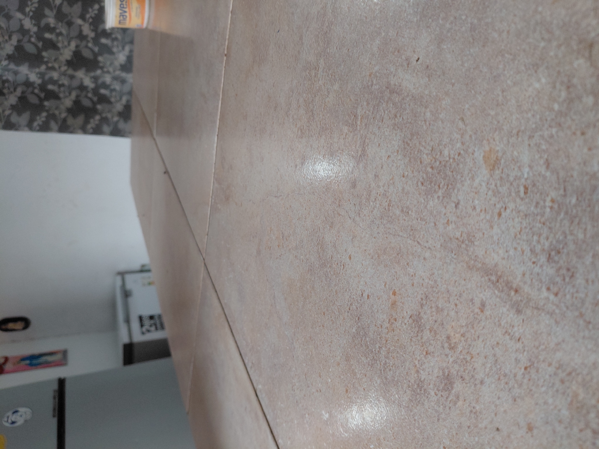 The image displays an indoor scene, primarily focusing on a tiled surface in the foreground and mid-ground, extending from bottom-right to top-left of the frame when viewed in its original orientation. The tiles are a mottled light brown or beige, featuring variations in shade, darker flecks, and some lighter, possibly worn, patches. Reflections of light are visible on the glossy surface of the tiles.
In the upper-left section of the image (when correctly oriented), a wall is visible, covered with a dark, patterned wallpaper featuring light-colored, possibly floral or leafy, designs. On a surface in front of this wall, an orange and white cylindrical container is present, with the brand name "navesa" clearly visible in white text on its label.
The background, largely out of focus, suggests a kitchen or utility room. Elements include a large, light-colored appliance, likely a refrigerator or freezer, with some items or stickers on its door, visible on the far left. Another white appliance or cabinet is partially visible further to the right. No people or specific activities are depicted. The scene appears to be under artificial indoor lighting, based on the reflections. The image itself is presented on its side.