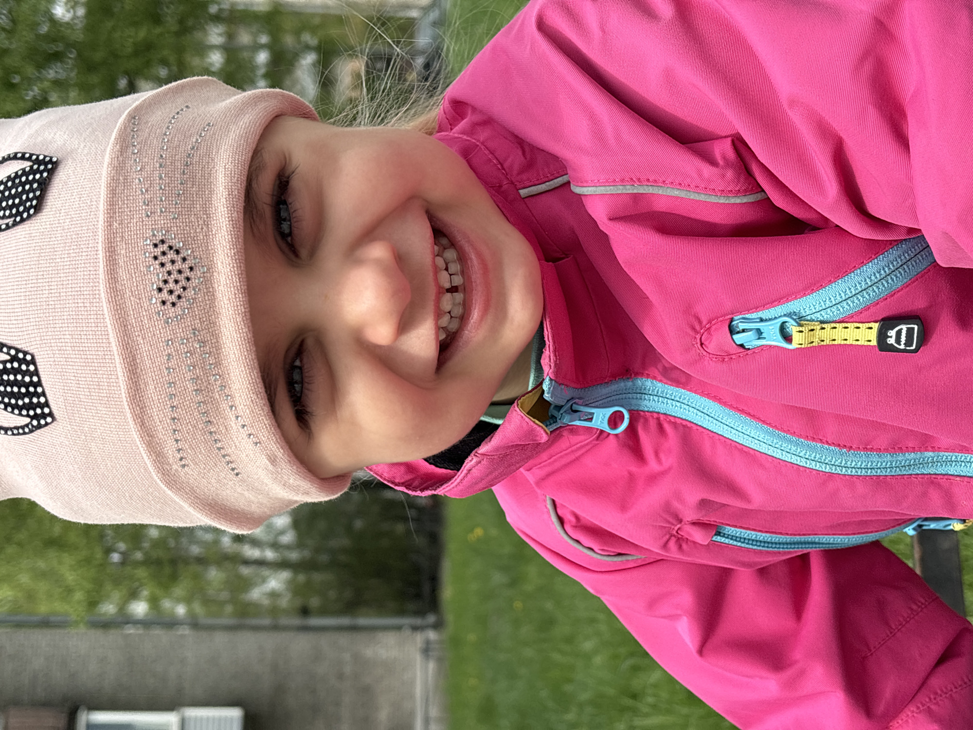 A young child with blonde hair is smiling at the camera. She is wearing a pink hat with decorative black and white spots and a pink jacket with blue zippers. The child appears to be outdoors in a grassy area. The location cannot be determined to be in the city of Saint Petersburg, Russia based on the information provided.