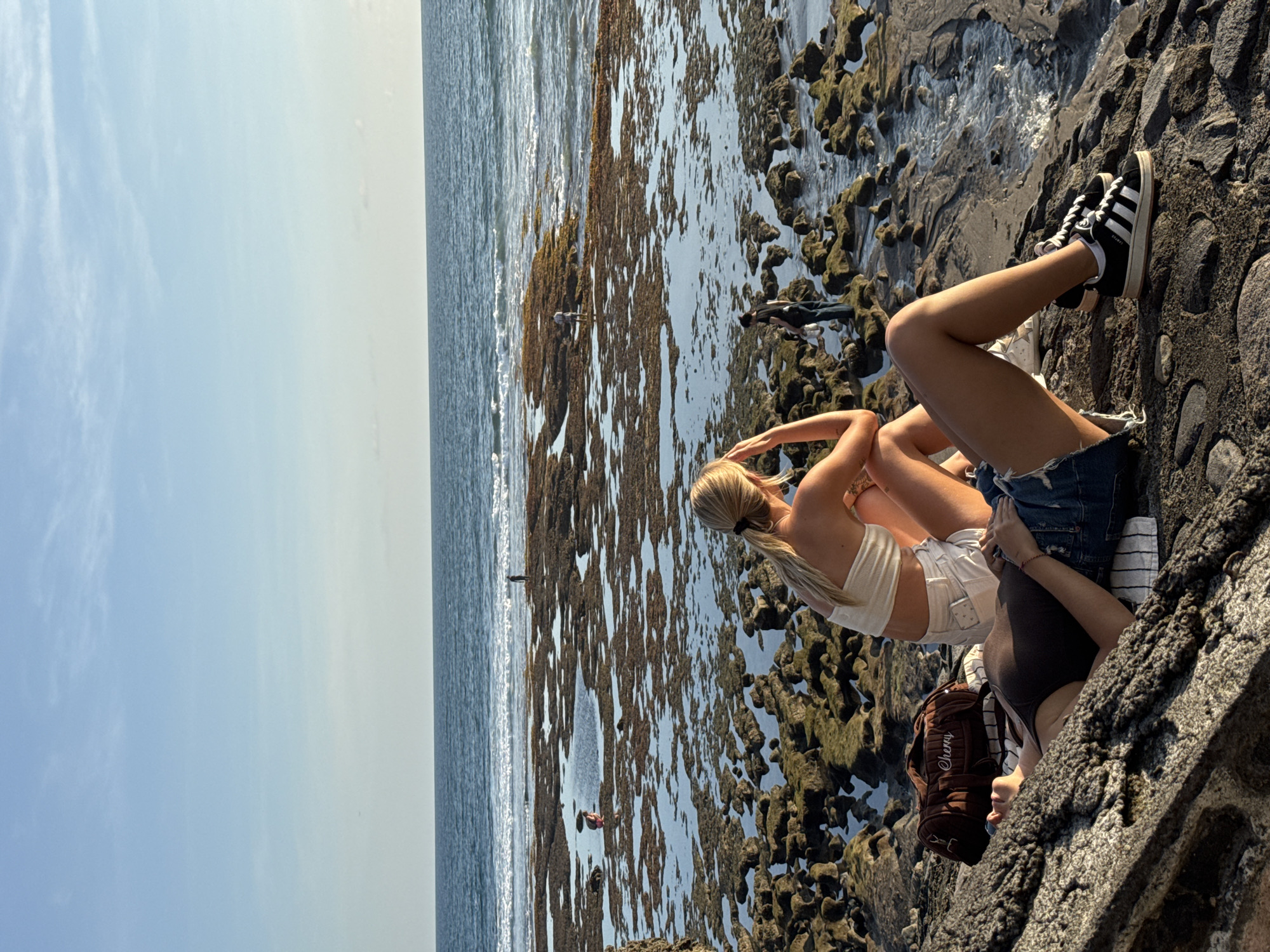The image captures a vibrant coastal scene in North Kuta, Indonesia, under a bright, clear sky. Two women are prominent in the foreground, seated on dark, rugged volcanic rock formations that extend into the sea.
One woman, with blonde hair tied in a ponytail, wears a white crop top and white shorts. Her arms are raised, hands resting behind her head, and her legs are crossed, suggesting a relaxed posture as she looks out towards the ocean. Beside her, the second woman, wearing a dark top and denim shorts, is seen with one leg bent and the other outstretched. She is wearing black Adidas Campus sneakers with white stripes and laces. A brown backpack with the word "Cherry" visible on it rests near her.
The midground features a vast expanse of dark, uneven rocks, heavily encrusted with dark green or brown marine growth, likely exposed during low tide. Patches of calm, reflective water are scattered amongst these formations. Several distant figures can be seen exploring these rocks, indicating activity further out on the tidal flats. Beyond the rocks, the deep blue ocean meets a clear horizon, under a bright, sunny sky with minimal, wispy clouds. The lighting suggests it is daytime, with strong natural light illuminating the scene. The overall atmosphere is one of leisure and appreciation of the natural coastal landscape.