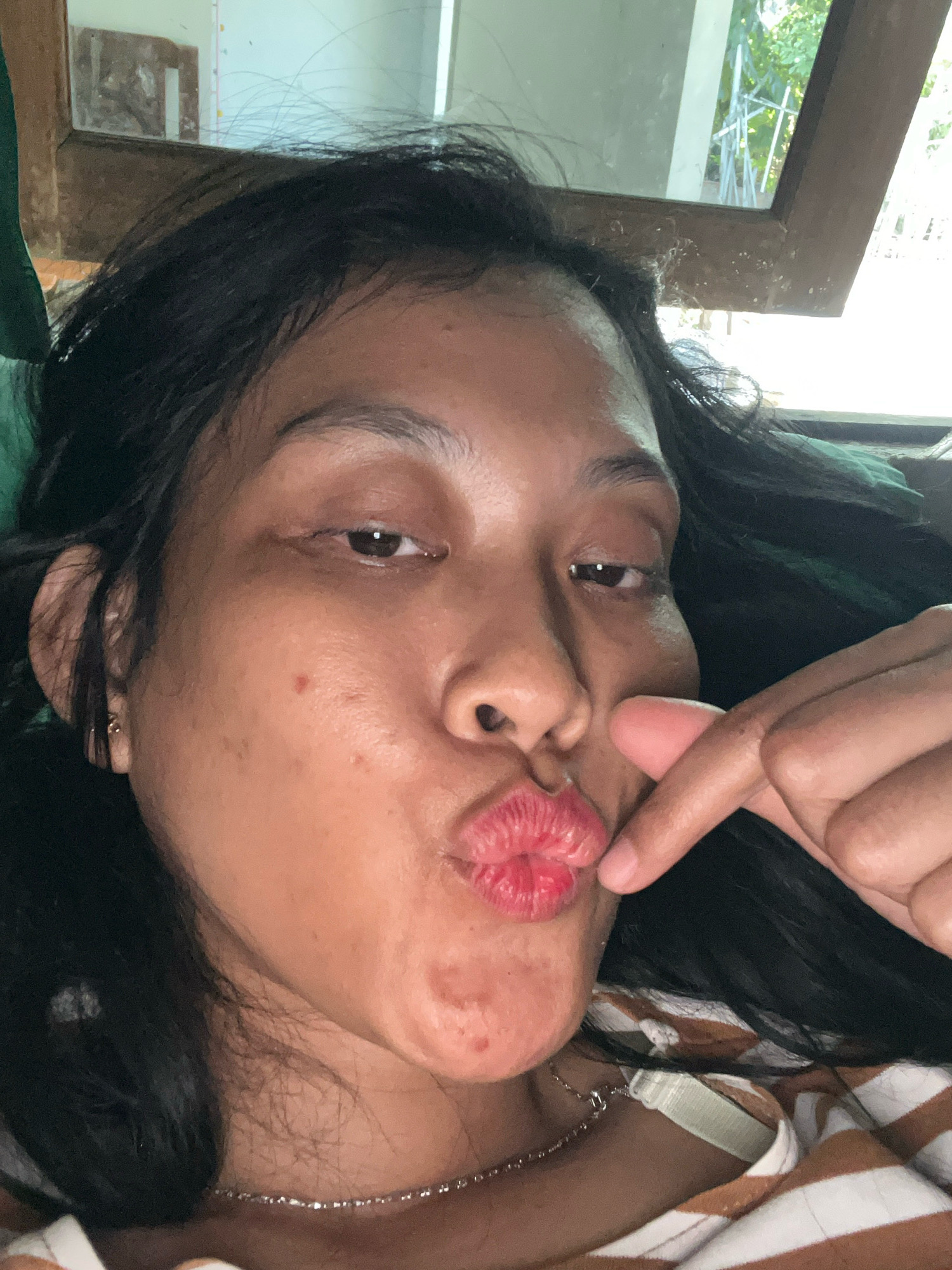 The media file is a close-up selfie of a person in Kecamatan Pati, Indonesia. The subject is a woman with dark hair, who is puckering her lips as if to give a kiss. Her right index finger is extended and touching her lower lip, creating a gesture that resembles a heart shape. She is wearing a striped shirt in shades of brown and white, and a delicate silver necklace. Her facial expression suggests playfulness or affection. The background is slightly blurred, but suggests an indoor setting with a window through which some greenery and possibly some construction materials are visible, indicating daylight. The overall mood is informal and cheerful.