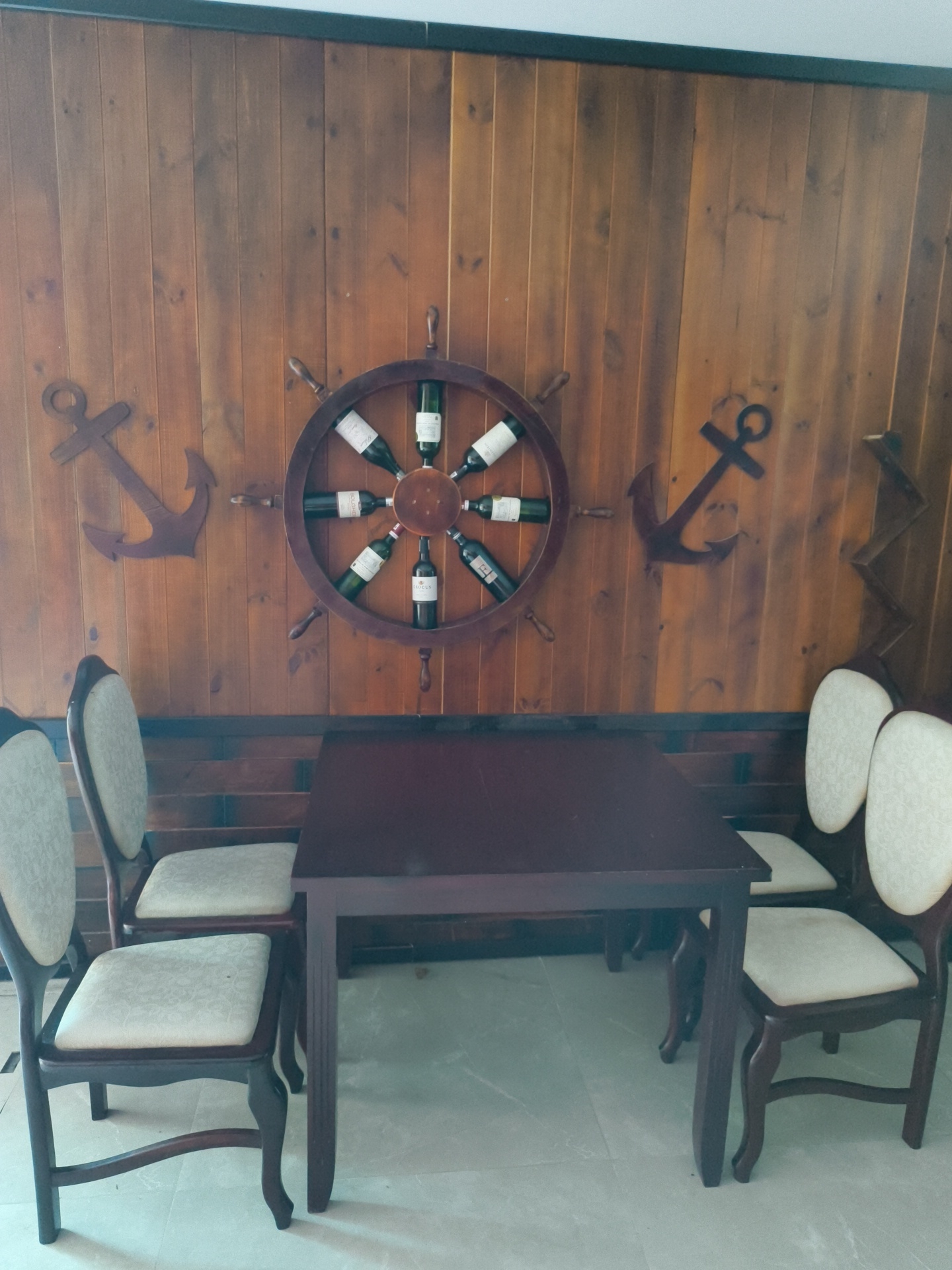 The image depicts a dining area with a nautical theme, situated in Phu Thuy Ward, Vietnam. The background is a wooden wall paneled with vertical planks. Centered on the wall is a wooden ship's wheel, acting as a wine rack, holding eight bottles of wine. On either side of the ship's wheel, two dark brown anchors are mounted on the wall. In the foreground, a dark brown rectangular table is positioned in front of the wall. Four chairs with light-colored, patterned upholstery are placed around the table; two are visible on the left side of the image, and two on the right. The floor appears to be tiled in a light gray color. The lighting is moderate, casting soft shadows. There are no people or animals present in the scene.
