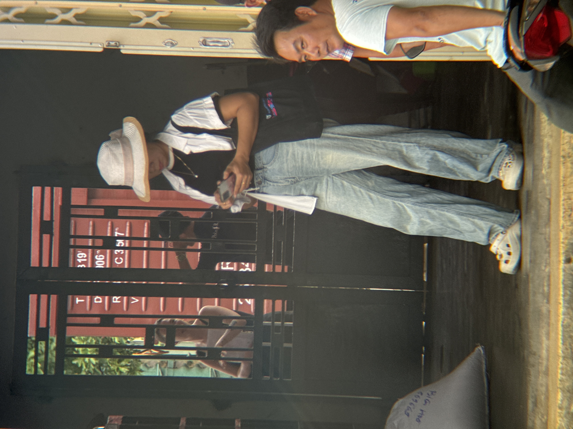 The image depicts an outdoor scene in Thuận Hóa Ward, Vietnam, with a focus on a woman standing near a gated entrance. The woman is wearing a wide-brimmed hat, a black top, a white open-collared shirt, and light-wash denim jeans. She is holding a mobile phone in her right hand and has a necklace with a small pendant. She is also carrying a black tote bag over her left shoulder. Her footwear appears to be white clogs.
Behind her, through the gate, a red shipping container with markings such as "T 319", "006m", and "C 3 5 7" is visible. Two other individuals are partially visible within the gated area; one woman in a white tank top, and another person in a dark shirt with "Polo R" visible on it.
To the right of the woman, a man in a light blue t-shirt is looking towards the camera. He is positioned on a motorcycle, the red taillight of which is visible in the lower right corner.
In the lower left of the image, a large grey sack with blue writing is partially visible. The environment appears to be a street or alleyway with a textured ground surface.