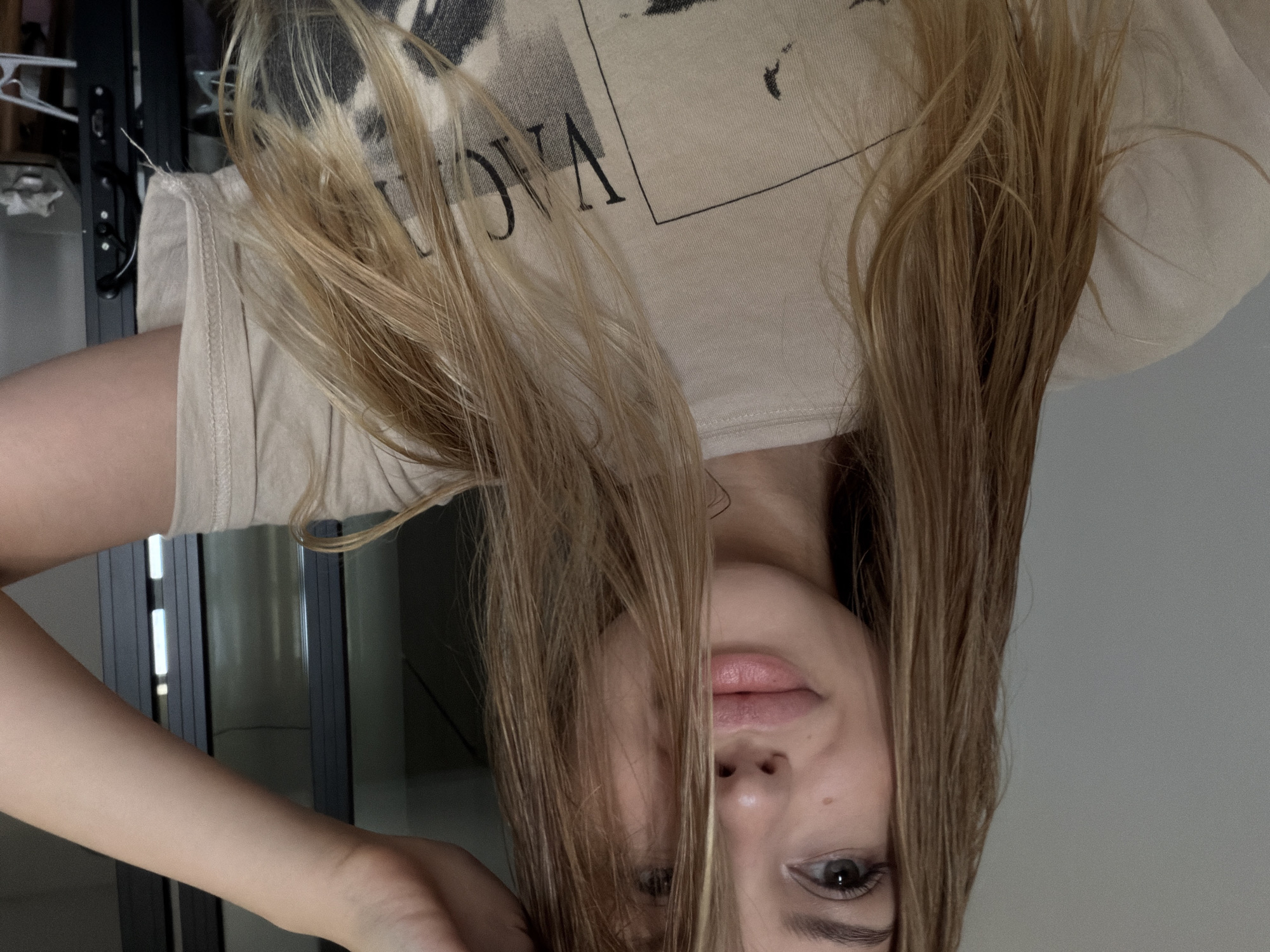 A young woman with blonde hair is taking a selfie indoors. She is wearing a light-colored t-shirt with a graphic print. Her left hand is raised to her head, and her hair is falling over her face. She is smiling and looking at the camera. The background is out of focus and appears to be a room with a window or glass door. There are no visible indicators of the city of Đà Nẵng, Vietnam.