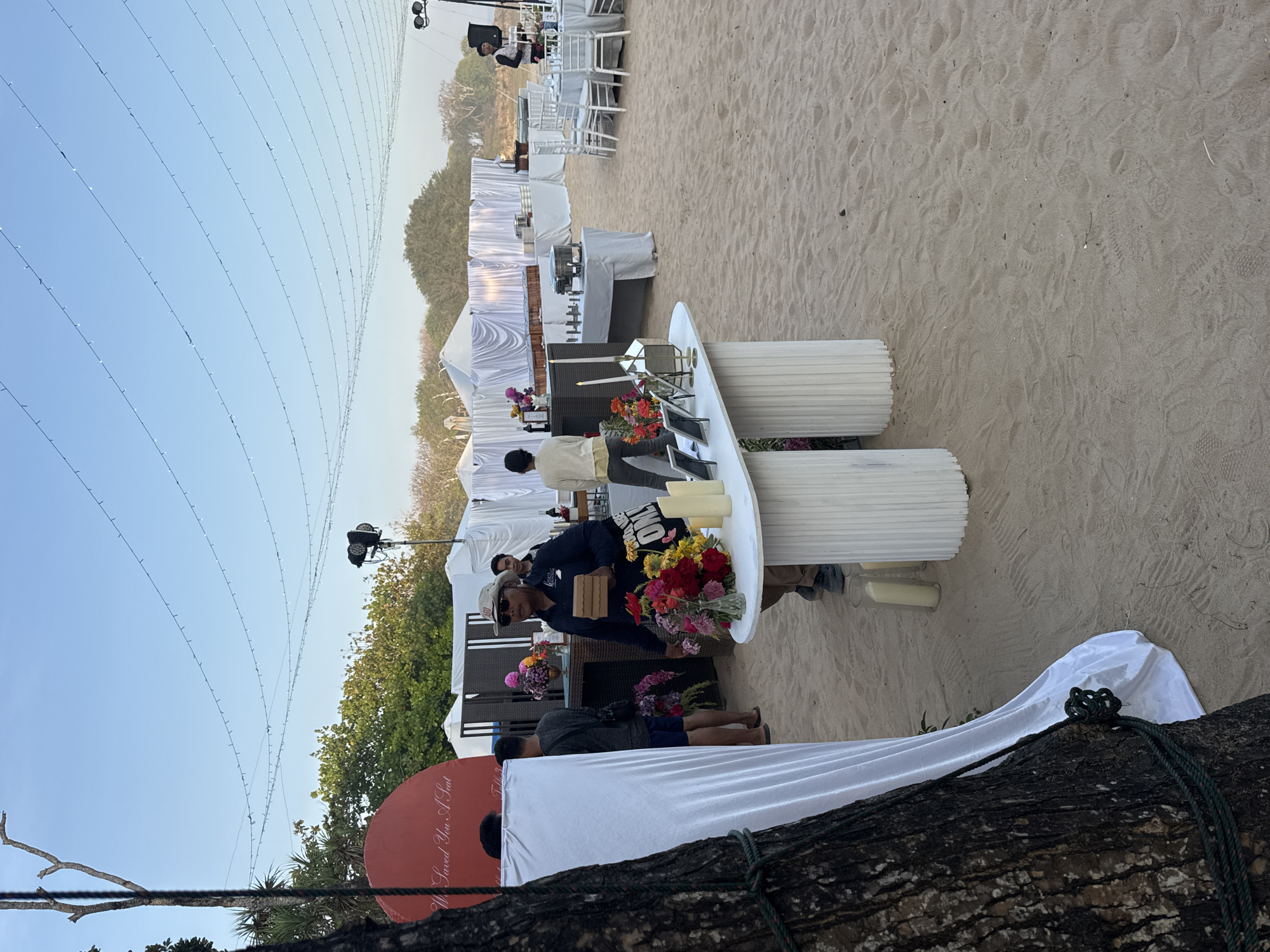 An outdoor event setup is observed on a sandy beach. Multiple white draped tents are visible in the background, with tables laden with food and decorations. In the foreground, a white, fluted table displays floral arrangements, candles, and framed photos. A man wearing a hat and sunglasses walks past, carrying a small box. Other individuals are seen milling around the setup. Strings of lights are strung across the sky above the tents. The scene appears to be in Kuta Selatan, Indonesia, likely set up for a wedding reception or a similar gathering on the beach.