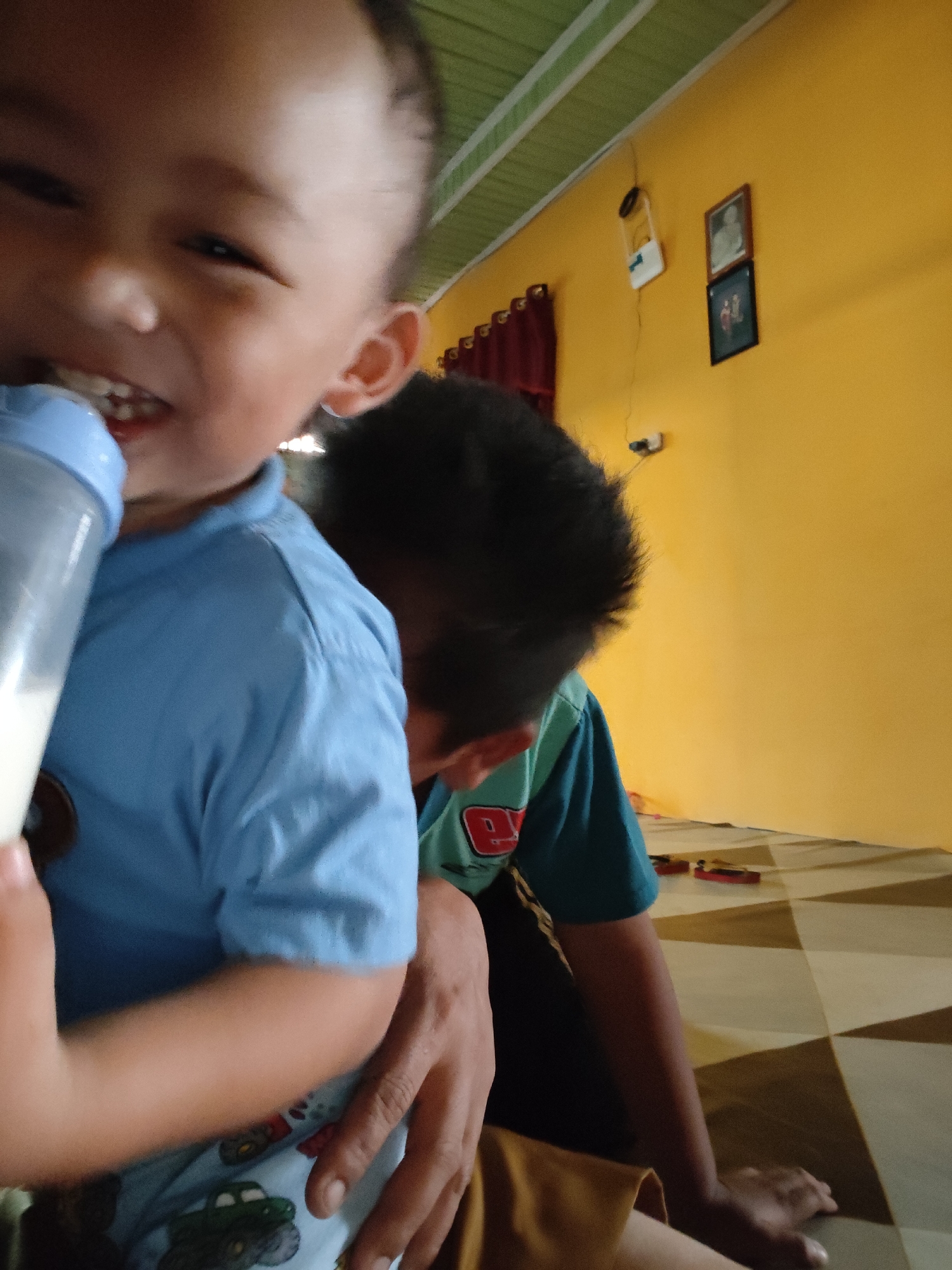 The image depicts a close-up view of two young children indoors, likely in a residential setting in Tanah Grogot, Indonesia. In the foreground, a baby in a light blue t-shirt is holding a milk bottle to their mouth. The baby's face is partially obscured but appears to be smiling. Behind the baby, another child with dark hair is visible, wearing a teal t-shirt with red lettering. This child is seated and leaning forward. The background features a yellow wall with two framed pictures and a dark red curtain. The floor has a tiled pattern. The overall scene suggests a casual, domestic moment.