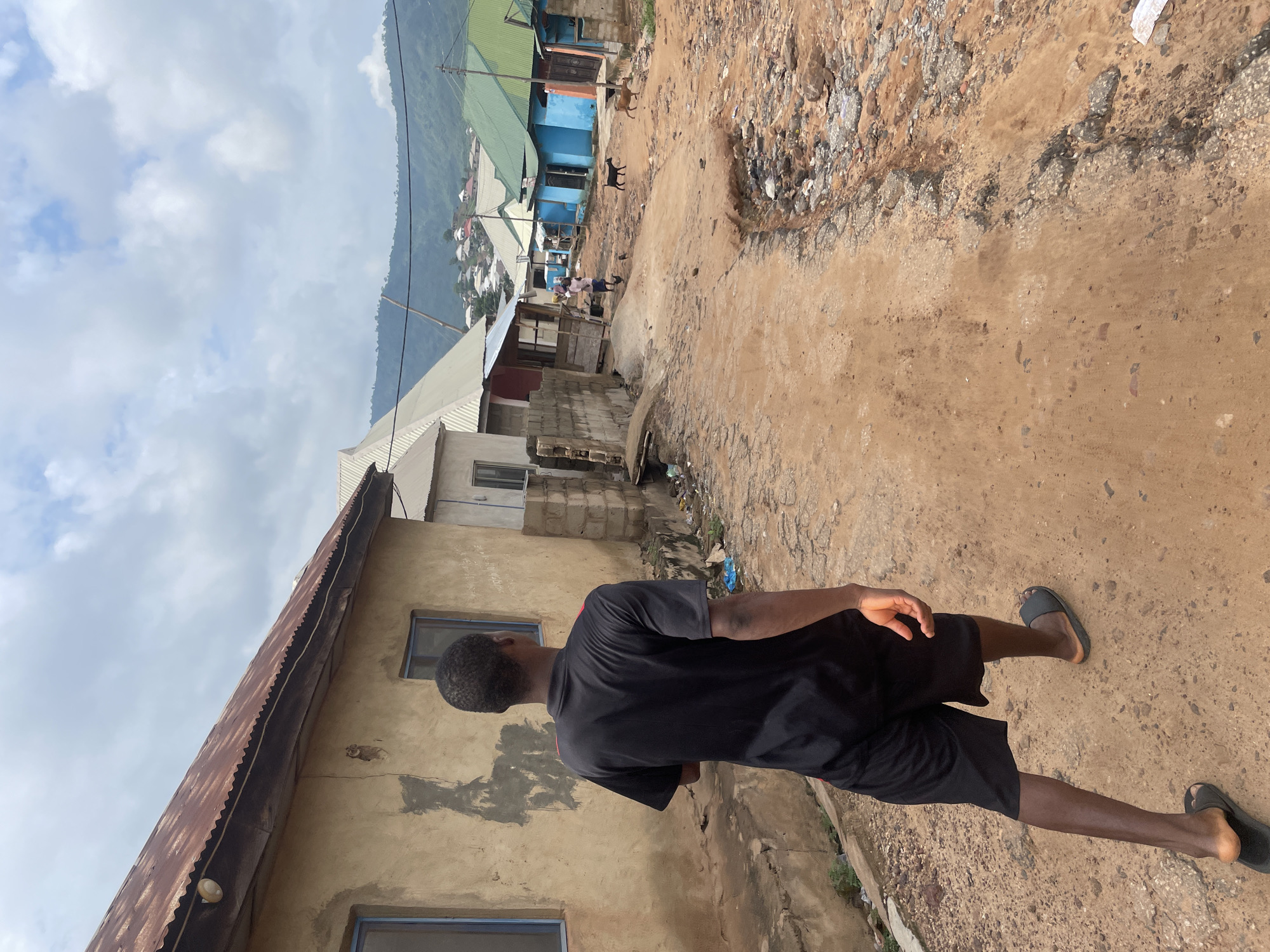 A young Black male wearing a black t-shirt and black shorts walks away from the camera on a dirt road. He is wearing black slide sandals. To his left is a dilapidated building with a rusty corrugated metal roof. The building's walls are a light tan color with some areas patched with gray cement. Blue framed windows are visible on the building. To the right of the road, several other structures are visible, some with corrugated metal roofs painted green and blue. In the distance, a hilly landscape with a town built on its slopes is visible. Two goats are visible on the road in the middle distance, and several chickens are near a person carrying a bundle on their head. The sky is cloudy. The overall scene suggests a rural or semi-urban environment in Lokoja, Nigeria.