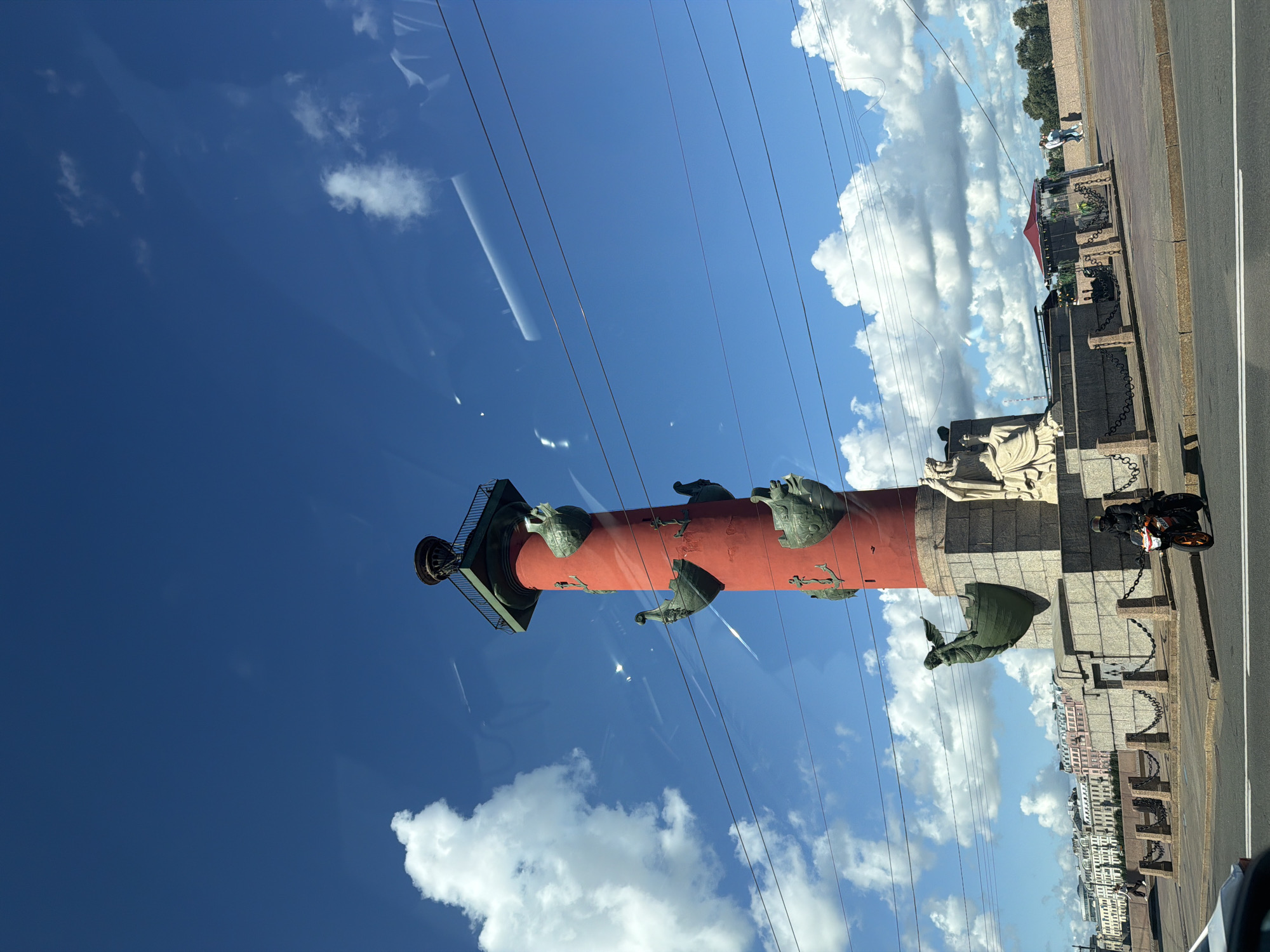 The image depicts the Rostral Columns, a prominent landmark in Saint Petersburg, Russia, situated on the spit of Vasilievsky Island. The Rostoal Column, painted a vibrant red, stands tall against a clear blue sky dotted with fluffy white clouds. Four ships' prows, each adorned with winged figures, jut out from the column's sides, and anchors are visible between them. The column is topped with a decorative lantern. At the base of the column, granite structures are adorned with sculptures of seated female figures. A motorcycle rider, clad in a black jacket and helmet, is positioned on the paved area in front of the column. In the background, across a body of water, is a row of colorful buildings, characteristic of Saint Petersburg's architecture. The scene is further characterized by a chain fence bordering the paved area and power lines stretching across the sky.
