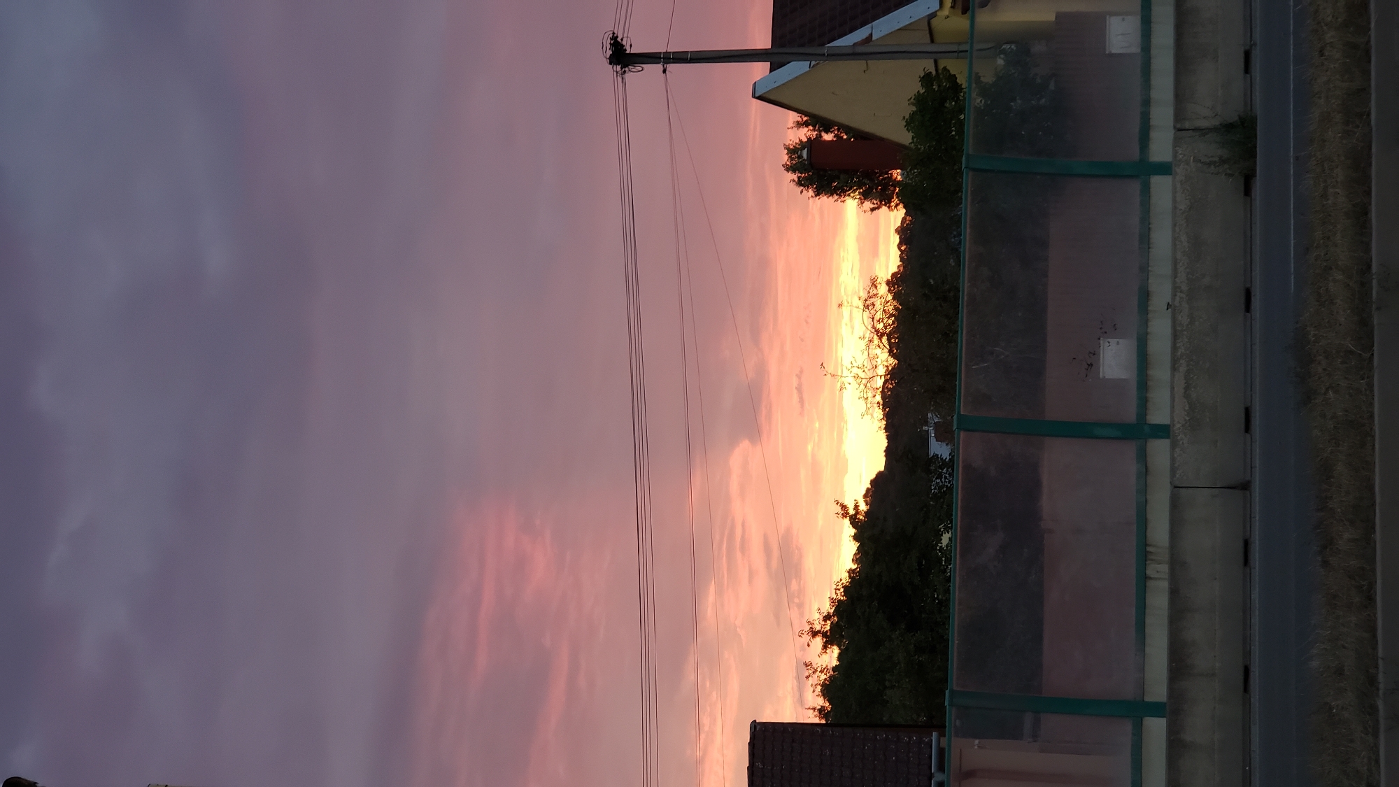 The image depicts a sunset scene in Prešov, Slovakia. The sky is filled with vibrant colors, ranging from deep purple at the top to bright orange and pink hues near the horizon. Several wispy clouds are scattered across the sky. In the middle ground, there is a line of trees, forming a dark silhouette against the colorful sky. Behind the trees, glimpses of houses and buildings can be seen, suggesting an urban or suburban environment. In the foreground, there is a barrier, possibly a sound barrier along a road, constructed with concrete blocks at the base and a translucent material with green framing above. Power lines are visible stretching horizontally across the upper part of the image, supported by a utility pole on the right side. The overall scene captures the beauty of a sunset over a town.