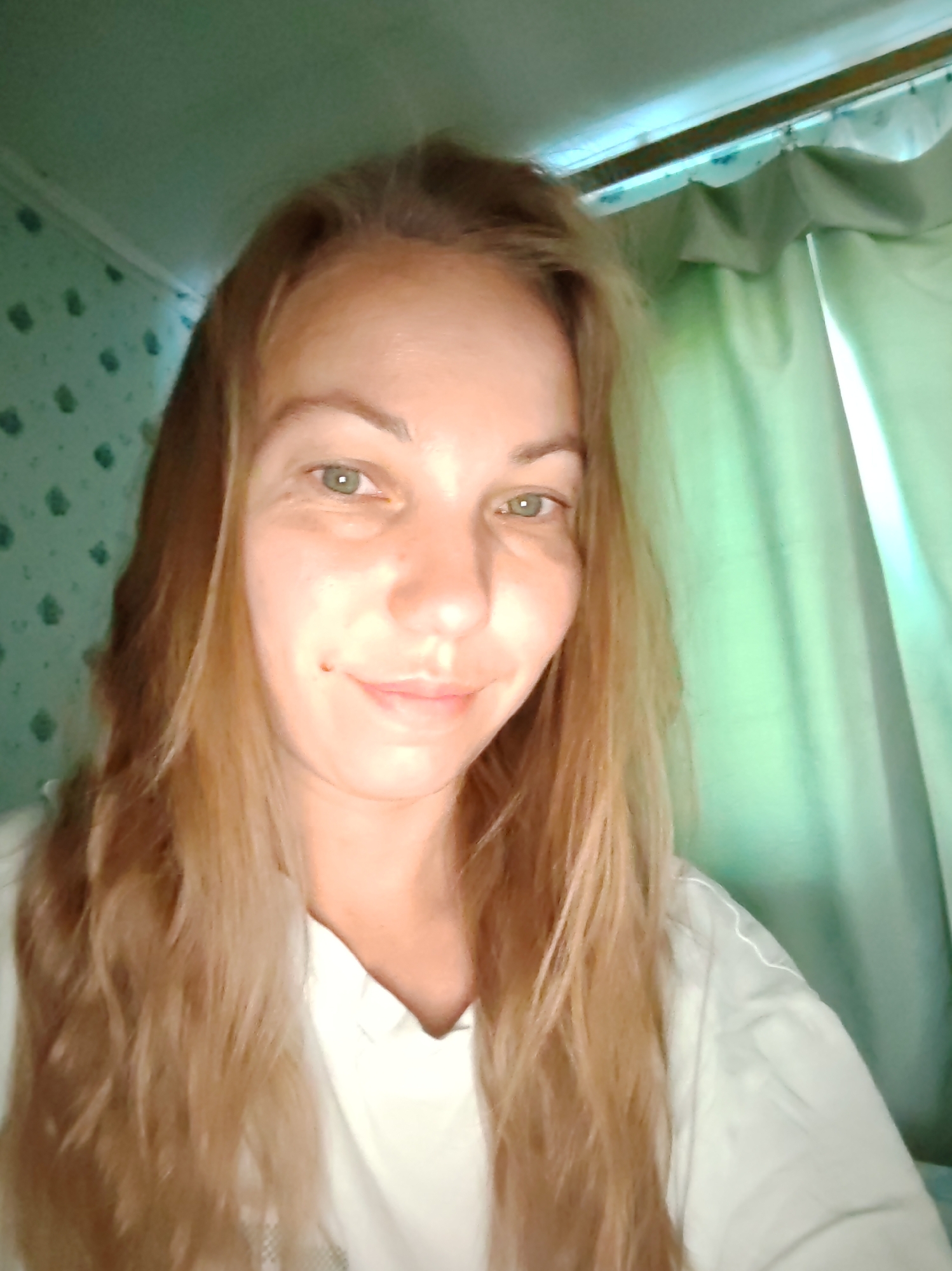 The image is a selfie taken by a woman with long, blonde hair. She is wearing a white shirt. Her eyes are green. She has a small mole on her right cheek. The background is a room with a patterned wallpaper on the left and a green curtain on the right. The lighting is dim and appears to be natural. The image does not contain any information that would allow for the identification of the location within the city of Чухлэм, Russia.