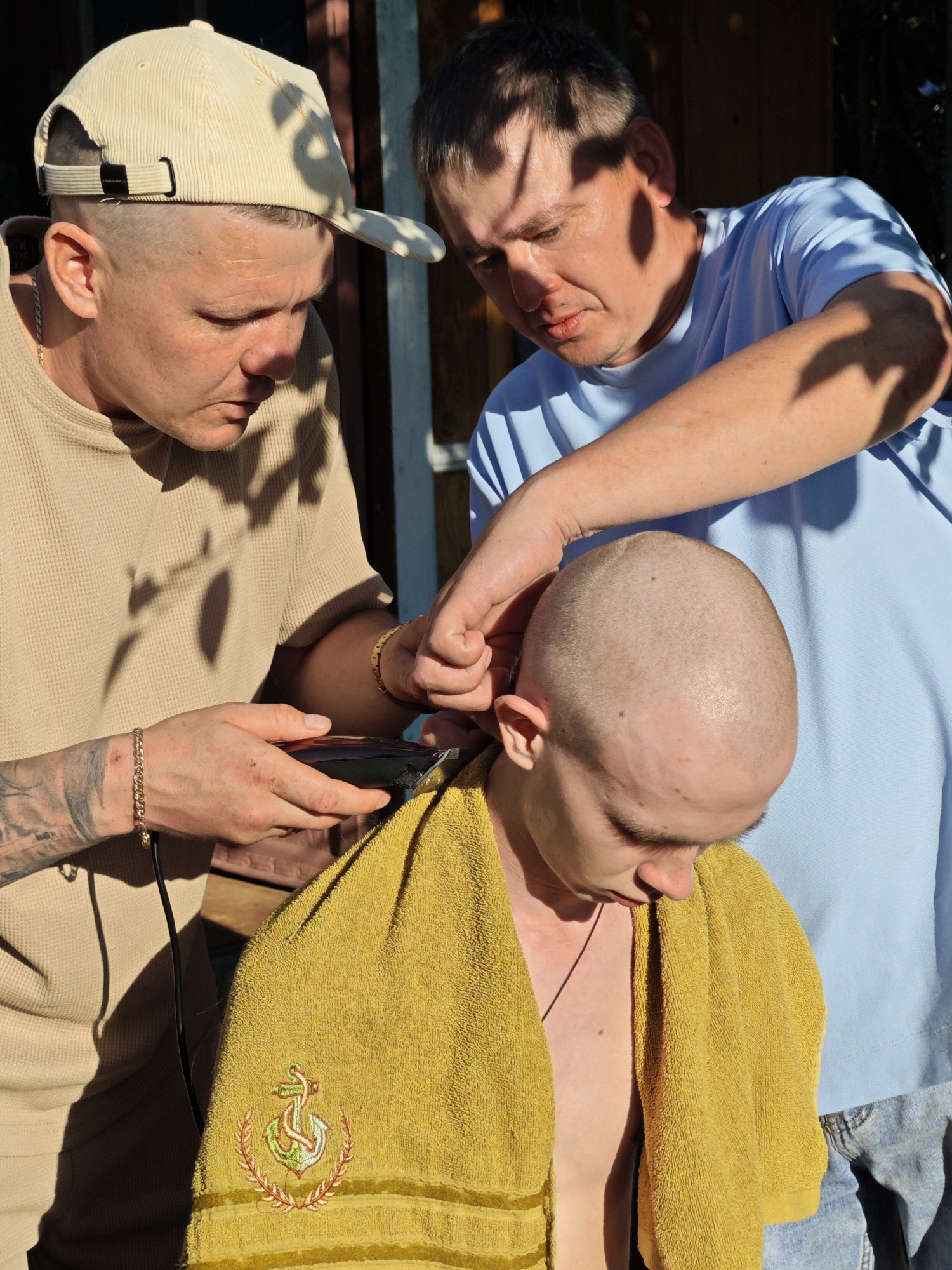 In the city of Foky, Russia, two men are outside, with one giving the other a haircut. The man receiving the haircut is wearing a yellow towel draped around his shoulders and has a buzz cut. The man giving the haircut is wearing a tan shirt and a beige baseball cap, and is holding an electric hair clipper. A third man, wearing a light blue t-shirt, is standing behind the man getting the haircut and appears to be assisting with the haircut by holding the person's head. The background suggests an outdoor setting, possibly a yard, with wooden structures and sunlight casting shadows.