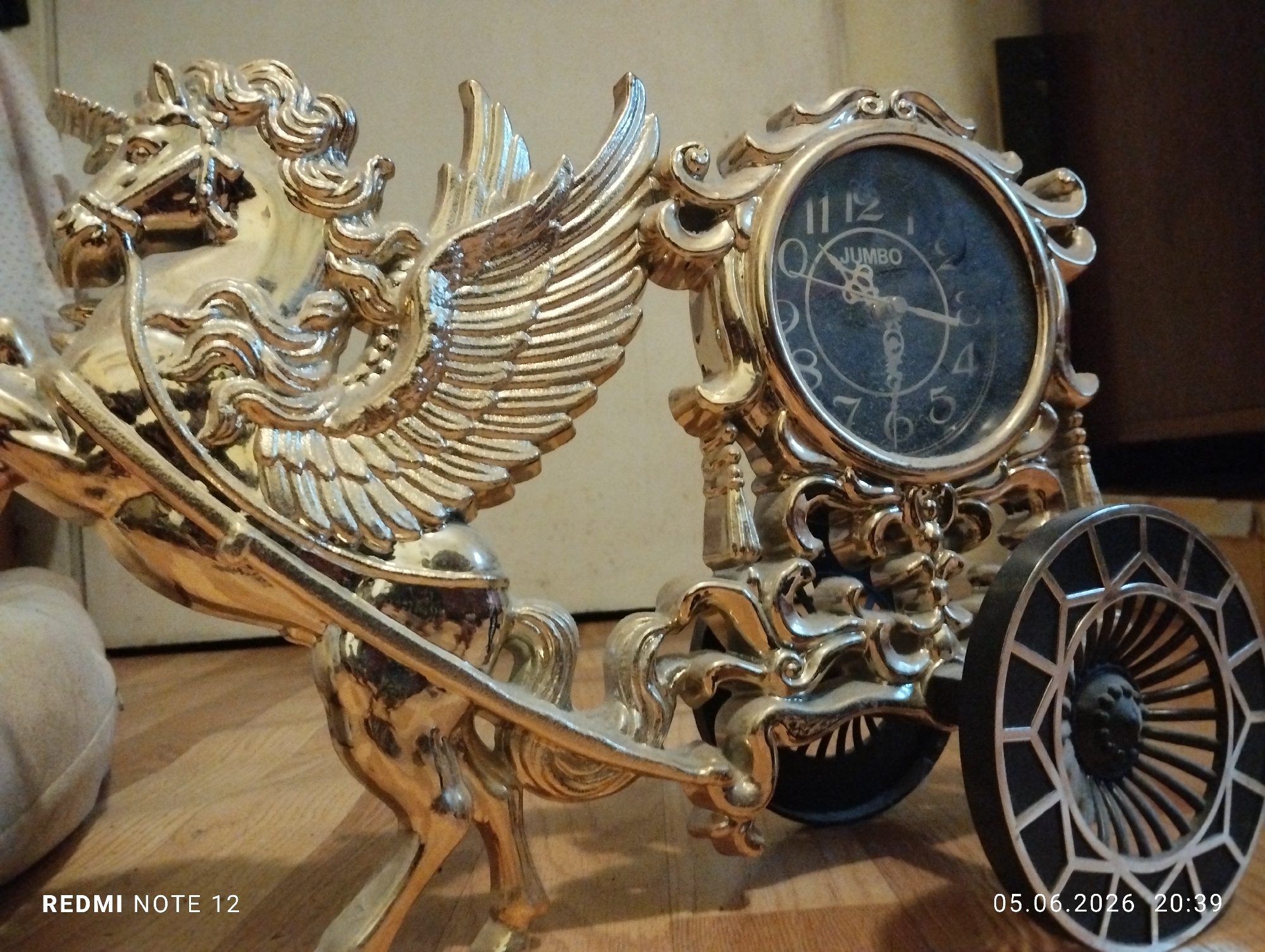 The scene is an interior located in the city of Burgas, Bulgaria. A gold-colored, ornate decorative clock, resembling a chariot pulled by a winged unicorn, is positioned on a light brown wooden plank floor. The unicorn figure, depicted in a rearing posture, features a detailed mane, tail, a bridle, a single horn on its forehead, and prominent wings extending backward. It is harnessed to an intricately detailed gold chariot frame that encases a round clock. The clock face is black with white Arabic numerals, indicating approximately 11:30, and the word "JUMBO" is printed above the center. The chariot's rear wheel is prominent, black with geometric silver-colored spoke details. The background to the left is a light, indistinct surface, possibly fabric, while the right background is a dark, undefined area. No individuals are present. The image includes embedded metadata indicating the camera model "REDMI NOTE 12" and a timestamp of "05.06.2026 20:39".