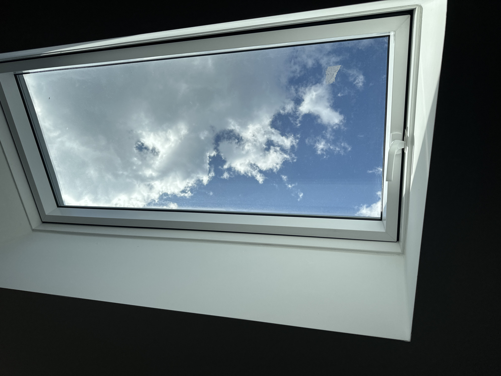 The image shows a view through a white-framed window looking up at a bright blue sky with scattered white clouds. The window frame has a white handle on the bottom edge. The interior of the room has dark walls and white trim around the window. The sun is casting a shadow across the white trim on the bottom of the window. The scene appears to be indoors. There is no indication of the location being in Garmisch-Partenkirchen, Germany, nor are there any people or specific actions depicted.