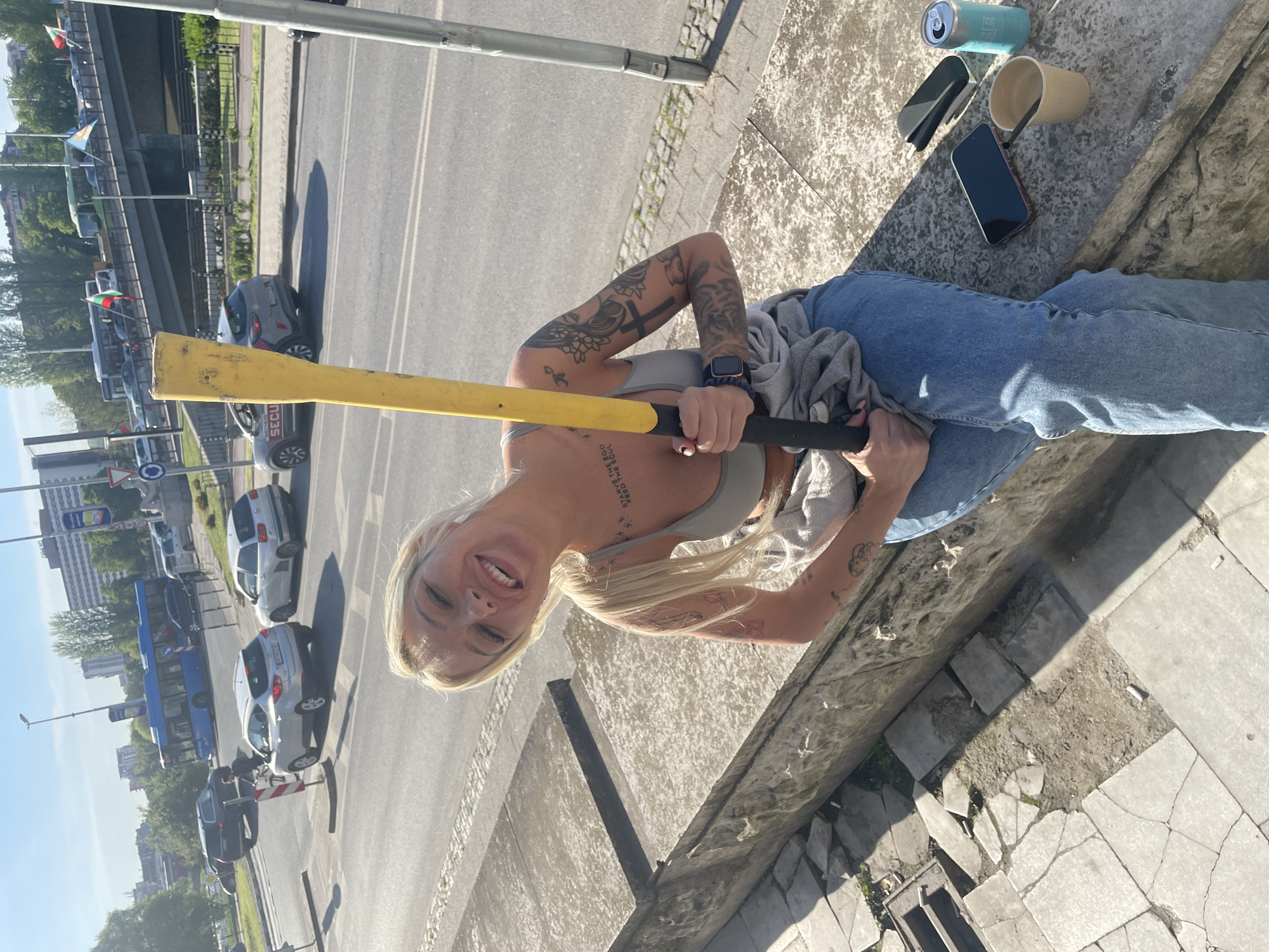 A young woman with blonde hair and numerous tattoos is captured in an outdoor setting on a sunny day. She is smiling brightly at the camera, holding a yellow and black object that resembles a bat or a tool handle. She is dressed in a grey sports bra and blue jeans, with a grey sweatshirt tied around her waist. A smartwatch is visible on her left wrist.
She appears to be sitting or leaning on a rough stone or concrete ledge. Beside her on the ledge are a light blue beverage can, a tan paper cup with a black straw, a black electronic device (possibly a vape or power bank), and a smartphone.
In the background, a busy street is visible with various vehicles including cars (grey, white, black) and a blue bus. One grey car has "SECU" visible on its side. European-style traffic signs, including a circular blue sign and a triangular red warning sign, line the road. Further in the background, flags displaying white, green, and red stripes are seen, indicative of Bulgarian flags, aligning with the Plovdiv, Bulgaria location context. Buildings and trees are also visible in the distance.
The bright daylight casts clear shadows, suggesting it is either mid-morning or afternoon. The ground near the ledge consists of cracked concrete tiles. Visible tattoos on the woman's arms include a prominent cross, floral patterns, and other designs. A chest tattoo is partially legible as "START THE...", "BE THE EGO...", "THE SOUL". The woman's smile conveys a cheerful emotion.