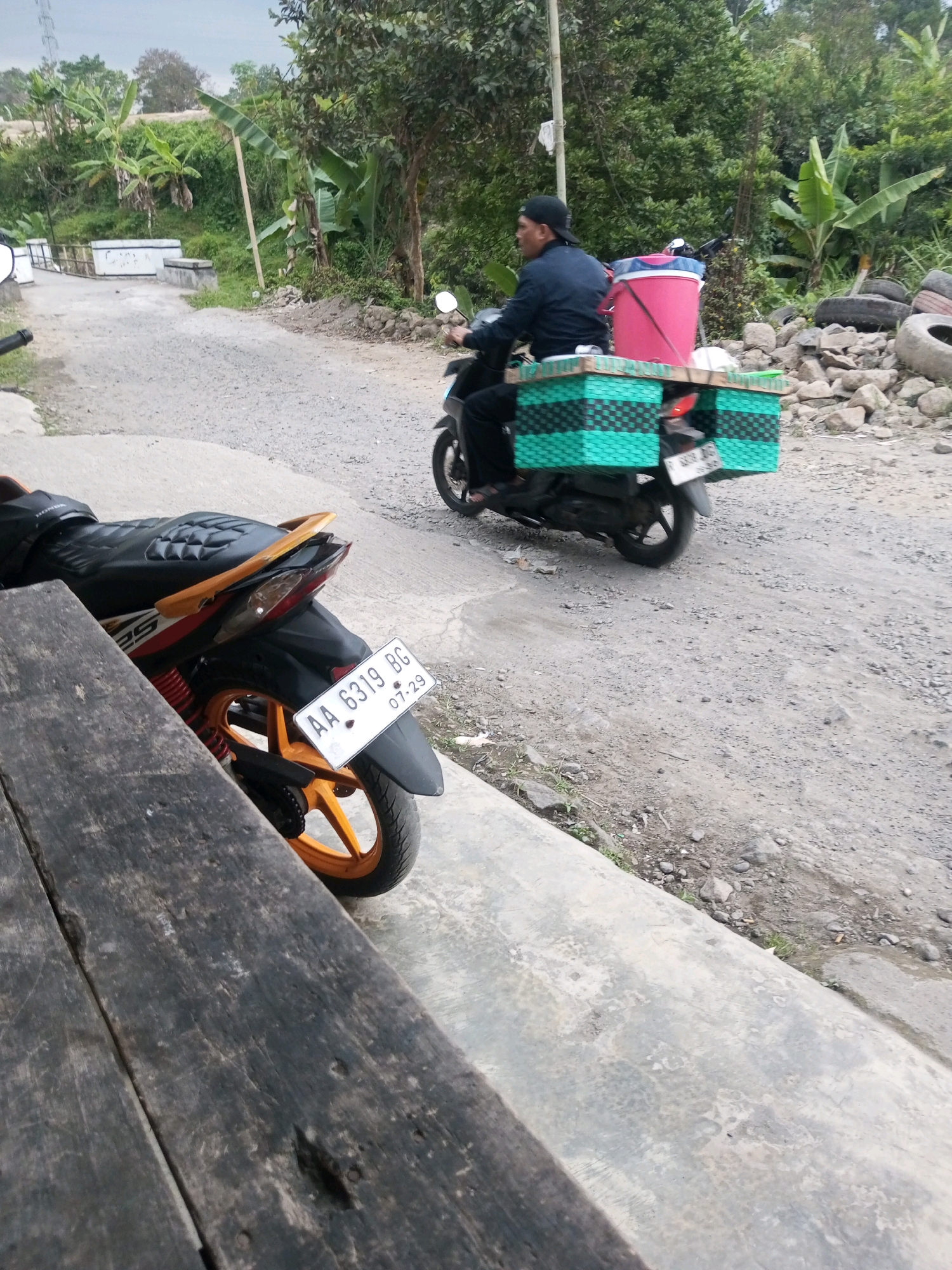 A man wearing a black cap and jacket rides a motorcycle with a pink bucket and two green woven boxes attached to the back. The motorcycle is on a dirt road, with trees and vegetation on either side. In the foreground, the rear of a parked motorcycle is visible, along with a wooden surface. The scene appears to be in Kalibeber, Indonesia, given the style of the motorcycle and the surrounding environment.