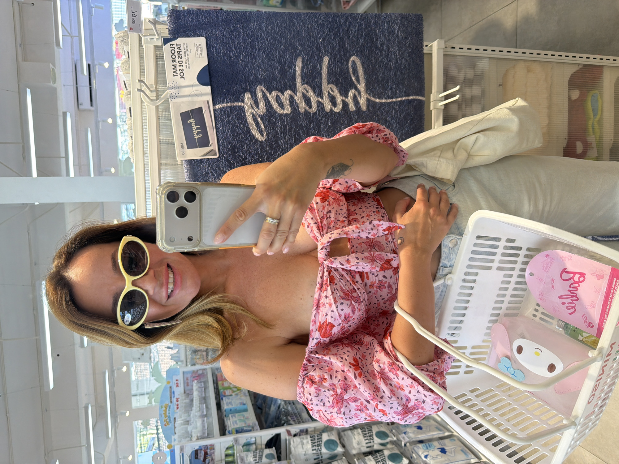 A woman with blonde hair and sunglasses is taking a mirror selfie in a store. She is wearing a pink floral top and blue jeans. She is holding a phone in her right hand and a shopping basket in her left arm. The basket contains a pink pouch with a cartoon character, a small green bottle, and a pink packet with text. Behind her, there is a blue mat with the word "goddess" written in white script. Shelves with various products are visible in the background, suggesting a retail environment. The lighting indicates it is daytime. The woman appears to be smiling and happy. The text "FLOOR MAT" is visible on a label attached to the mat. There is also text on the pink packet in the basket.