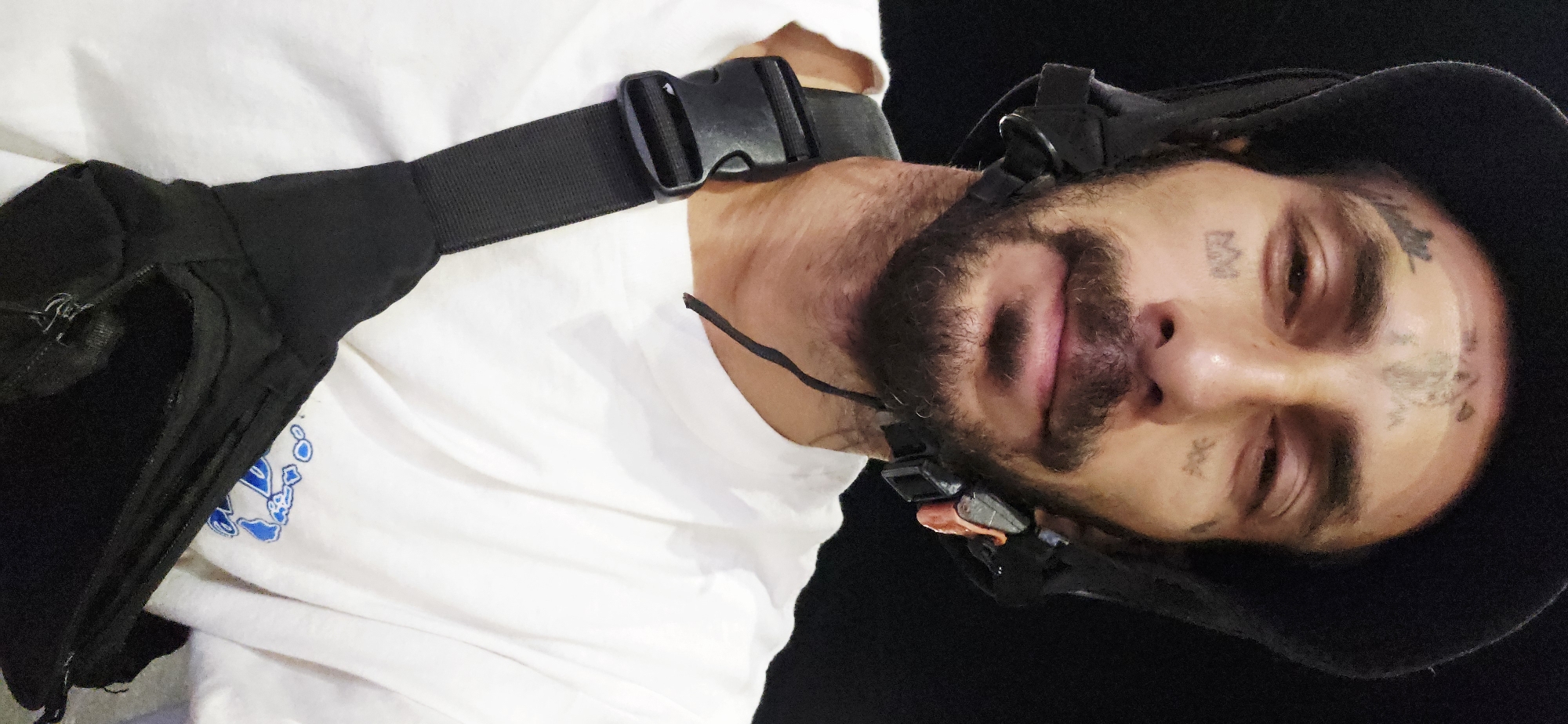 The image shows a close-up, upward-angled portrait of a man, possibly taken indoors or at night due to the dark background. The man is looking directly at the camera with a neutral expression.
He has a dark beard and mustache, and multiple tattoos are visible on his face. On his forehead, there's a symbol that looks like an "M" or a stylized crown, along with other faint script or symbols. More indistinct symbols are tattooed on his cheeks/temples. His eyes appear somewhat tired.
The man is wearing a plain white t-shirt with a partially visible blue graphic on the left side of his chest. Across his chest and shoulder, he has a black strap with a plastic buckle, which is part of a black fanny pack or sling bag worn across his body. A zipper of the bag is visible. He is also wearing a black hat or hood, and a black head-mounted device, possibly a VR headset or communication gear, is strapped around his head, with small orange and black components visible near his ear and mouth area.
The overall scene is a headshot of the man, focusing on his face and upper body against a dark, featureless background. No specific location cues are visible in the image itself, beyond the provided context of Cam Đức, Vietnam.