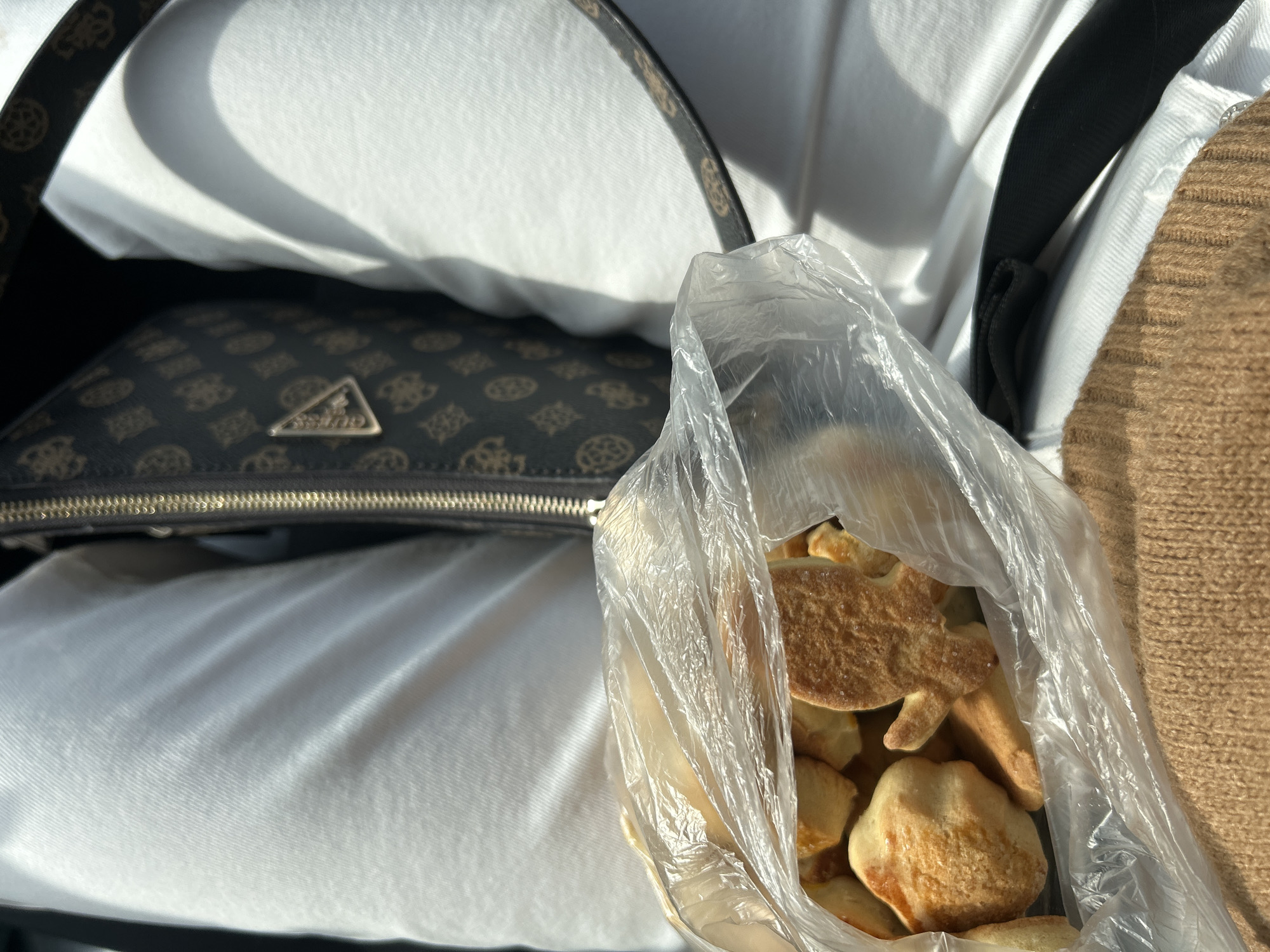 The image shows a partially filled, transparent plastic bag of baked goods, possibly cookies or pastries, held against a backdrop of white and brown fabrics. To the left, a brown Guess brand handbag with a logo pattern and a gold-colored zipper is visible. The lighting suggests the image was taken indoors, with soft natural light illuminating the scene. There is no indication or evidence suggesting the location is Церово, Bulgaria.