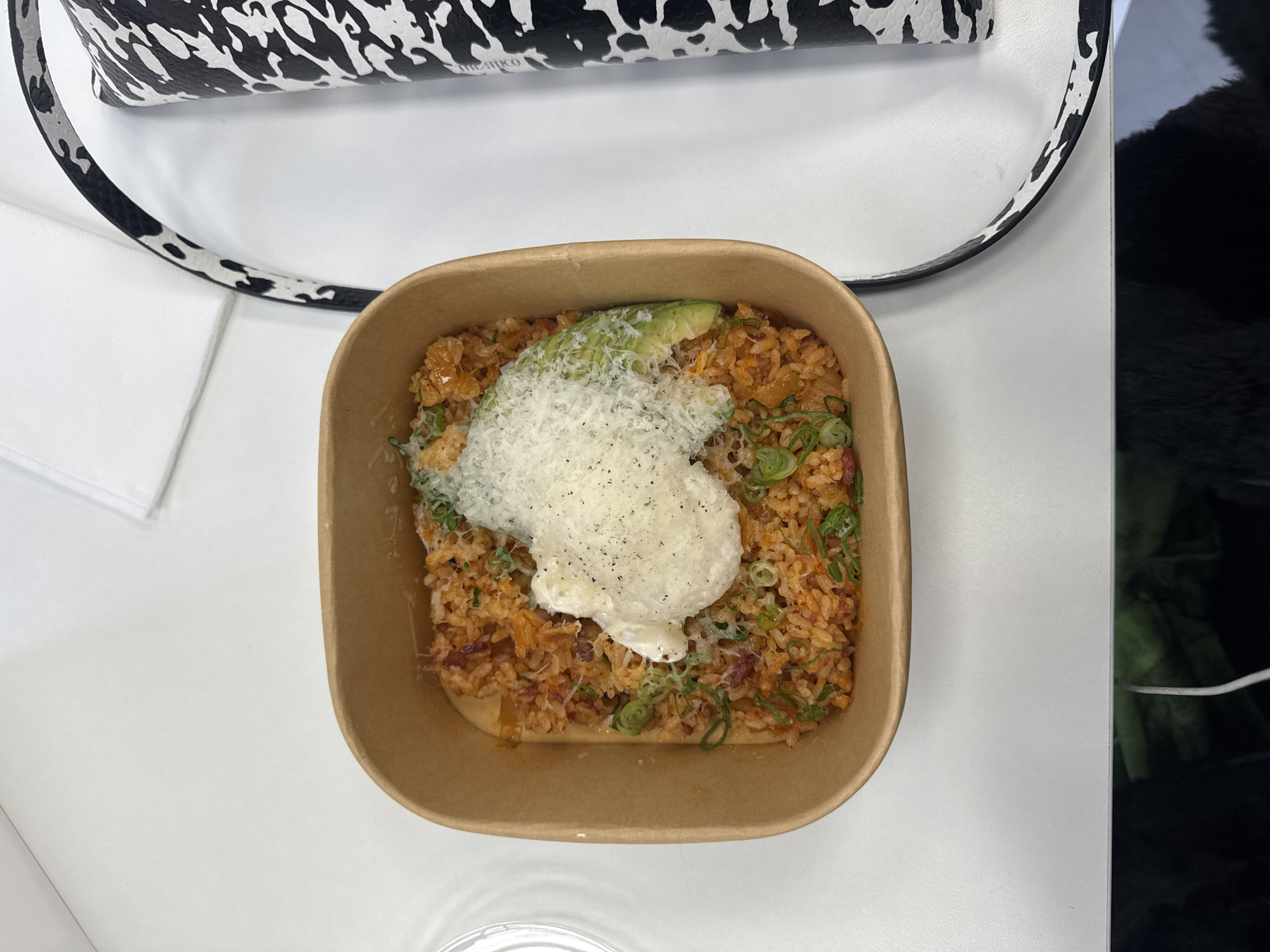 The image presents a top-down view of a takeaway container filled with food. The food appears to be a rice-based dish, possibly kimchi fried rice, topped with what seems to be a dollop of cream or sauce, grated cheese, and sliced avocado. Chopped green onions are scattered throughout the dish. The container is made of brown cardboard.
The background includes parts of a black and white patterned bag, a white napkin, a white table, and what appears to be a black object, potentially part of clothing or another item on the table.
There is no information available to determine the location of the image in Paris, France.