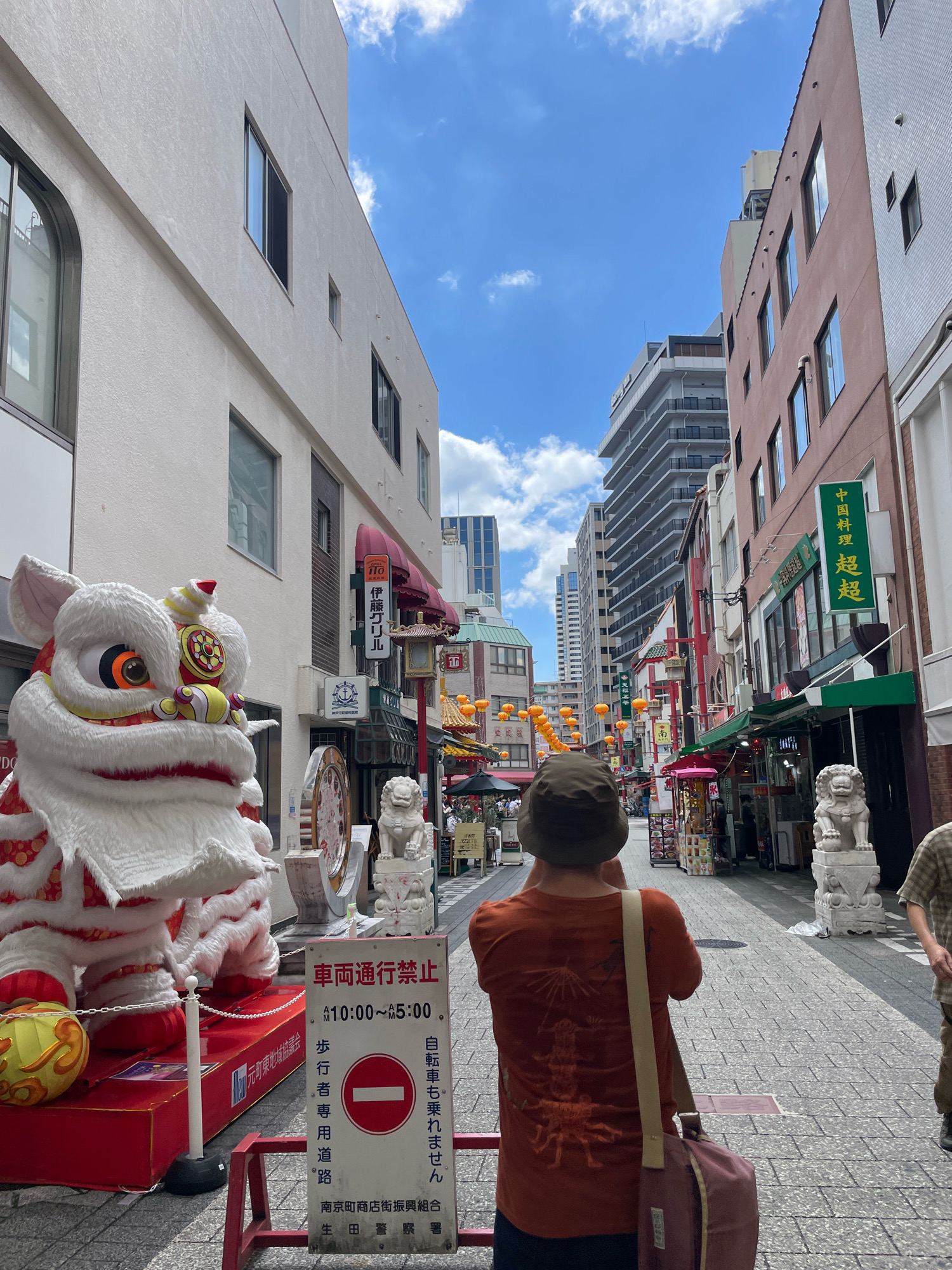 This is a daytime, eye-level, outdoor shot of a street in Kobe, Japan's Chinatown (Nankinmachi). In the foreground, a person wearing a tan hat and an orange t-shirt with a graphic design on the back is facing away from the camera, holding a smartphone and appearing to take a picture. A messenger bag is slung over their shoulder. To the left, a large, white, decorative lion statue stands on a red platform, decorated with red and gold accents, and a yellow lantern rests near its base. A red and white sign with Japanese text and a red circle with a horizontal bar is placed in front of the lion. The street is paved with gray cobblestones. To the right, a smaller white lion statue is visible, positioned on a pedestal. The street is lined with multi-story buildings, many of which have awnings and signs with Chinese characters. Orange lanterns are strung across the street. The sky is blue with scattered white clouds. The scene appears to be a busy tourist area.
