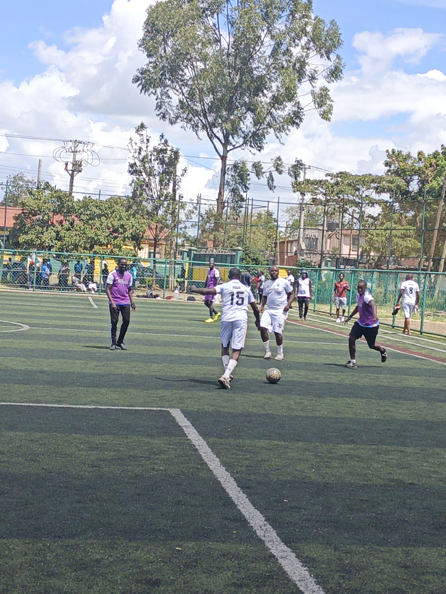 A daytime scene shows a soccer match underway on a green artificial turf field. Several players are visible, wearing different colored jerseys: white with purple accents and purple with white accents. One player wearing a white jersey with the number 15 is kicking the soccer ball. Other players are in various positions, jogging or running. Spectators can be seen behind a wire fence along the sidelines. The background includes trees, buildings, and utility poles with electrical lines. The sky is blue with scattered white clouds. The location is in Nairobi, Kenya, as indicated by the architecture.