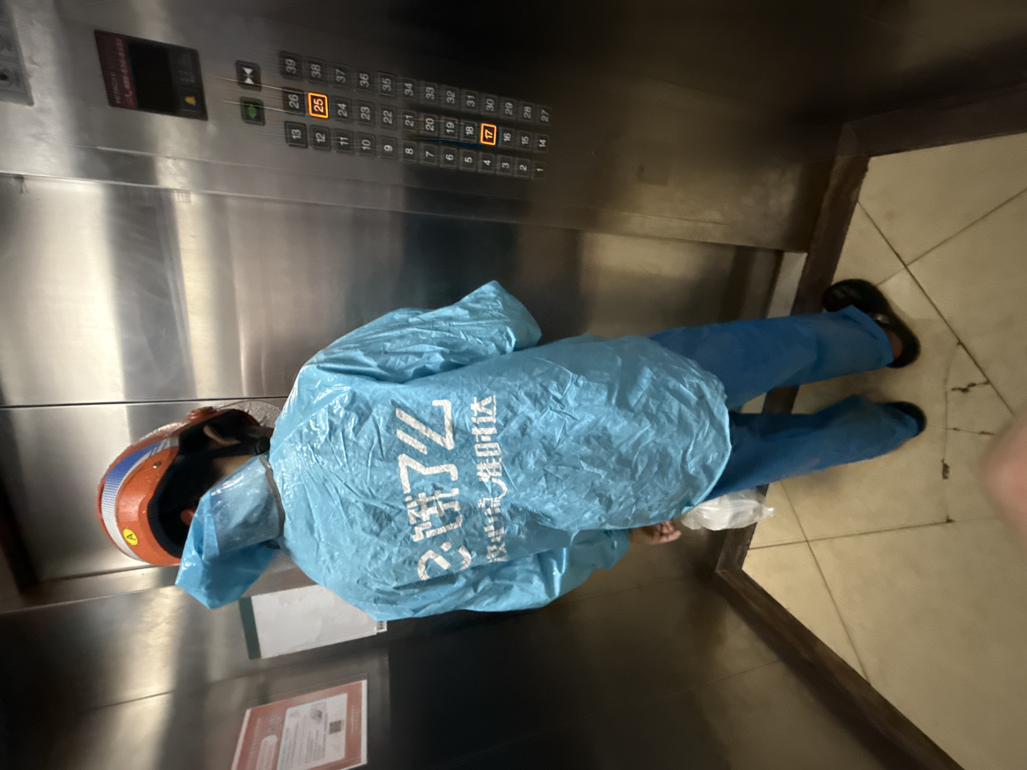 The image shows a person wearing a blue disposable suit and an orange helmet standing inside an elevator. The elevator has a stainless steel interior and a panel of illuminated buttons. The person appears to be a delivery person, possibly for "饿了么" (Ele.me), a food delivery platform, as suggested by the text visible on the suit. The text "饿了么 农业电流准时达" translates to "Ele.me, Agricultural electricity on time delivery". The elevator buttons show floor numbers, with floor 25 and 17 illuminated, indicating they are currently active. The scene appears to be indoors, likely in a residential or commercial building. The overall impression is of a routine moment in a delivery person's workday.