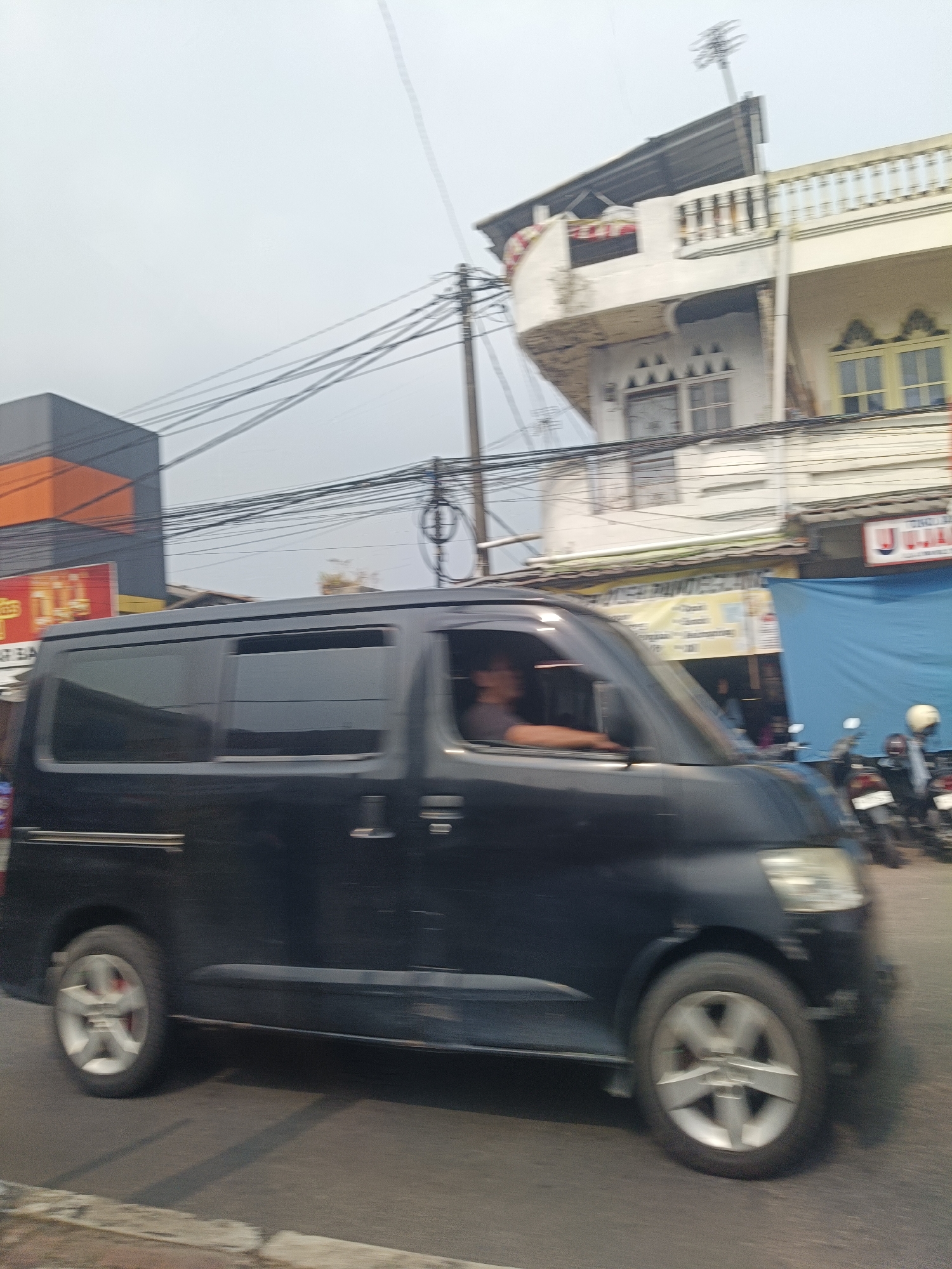 The image shows a black minivan in motion on a street in Pandeglang, Indonesia. The minivan is the primary subject, with its side profile visible as it travels from left to right. A person is seated in the driver's seat, with their arm resting on the open window. The background consists of buildings and power lines, suggesting an urban or semi-urban environment. One building, visible on the right, is a multi-story structure with balconies and decorative elements. A blue tarp is stretched across the front of one of the buildings. Several motorcycles are parked on the side of the street behind the blue tarp. The overall impression is of a bustling street scene, possibly in a commercial area. The sky is overcast.