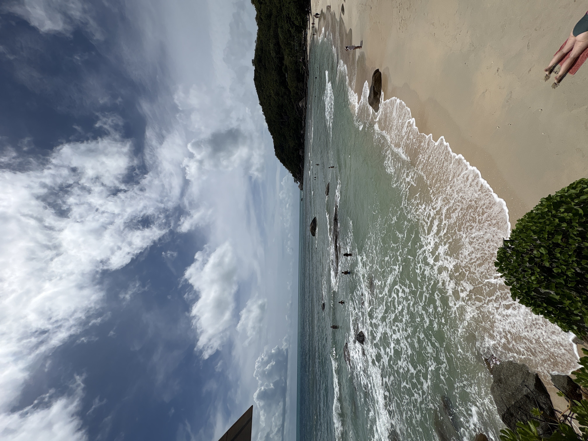 Baan Chaweng Noi, Thailand: Coastal scene with a sandy beach, clear turquoise water, and a lush green headland. People are swimming and wading in the water. The sky is partially cloudy with blue patches. A person's legs are visible on the sand. The ocean has visible waves. Vegetation borders the beach.