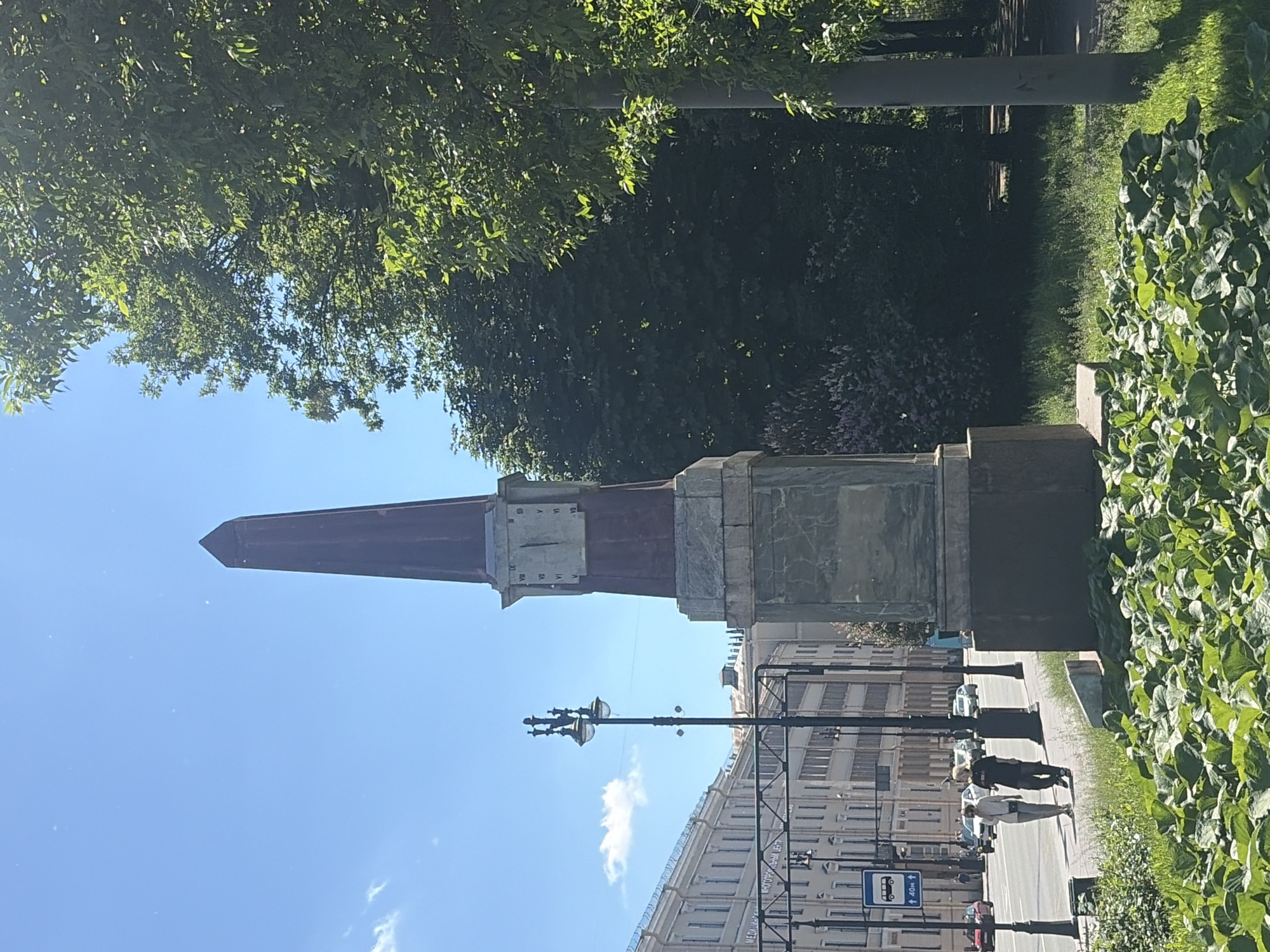 The image depicts an obelisk with a clock face, located in a park area of Saint Petersburg, Russia. The obelisk is made of stone with a reddish-brown, pointed top. The clock face on the obelisk shows Roman numerals. In the background, there is a street with buildings, a lamppost, and a bus stop sign. Two people are walking on the sidewalk. Lush green foliage and trees surround the obelisk, and the sky is clear blue with a few scattered white clouds. The scene suggests a sunny day in an urban park.
