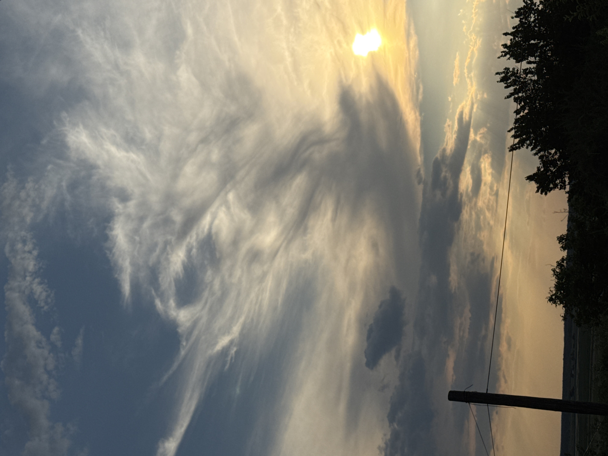 This is a vertical, low-angle shot of a sky at sunset. The sun is a bright, luminous white orb partially obscured by wispy, white clouds in the upper right quadrant of the image. These clouds are spread out and appear thin, catching the sunlight and glowing yellow and white. They are interspersed with darker, more substantial gray clouds that create shadows and add depth to the sky. In the lower right of the frame, the silhouette of a tall, dark tree with dense foliage is visible, its branches reaching towards the sky. A utility pole, also silhouetted, stands in the foreground, with a dark wire running diagonally across the image. The background appears to be a landscape, with hints of fields and possibly distant hills, rendered in muted tones of orange and yellow due to the sunset. The overall mood is serene and possibly a bit dramatic, with the contrast between the bright sun, the dark silhouettes, and the varied cloud formations. The time of day is clearly sunset. No text is visible in the image.