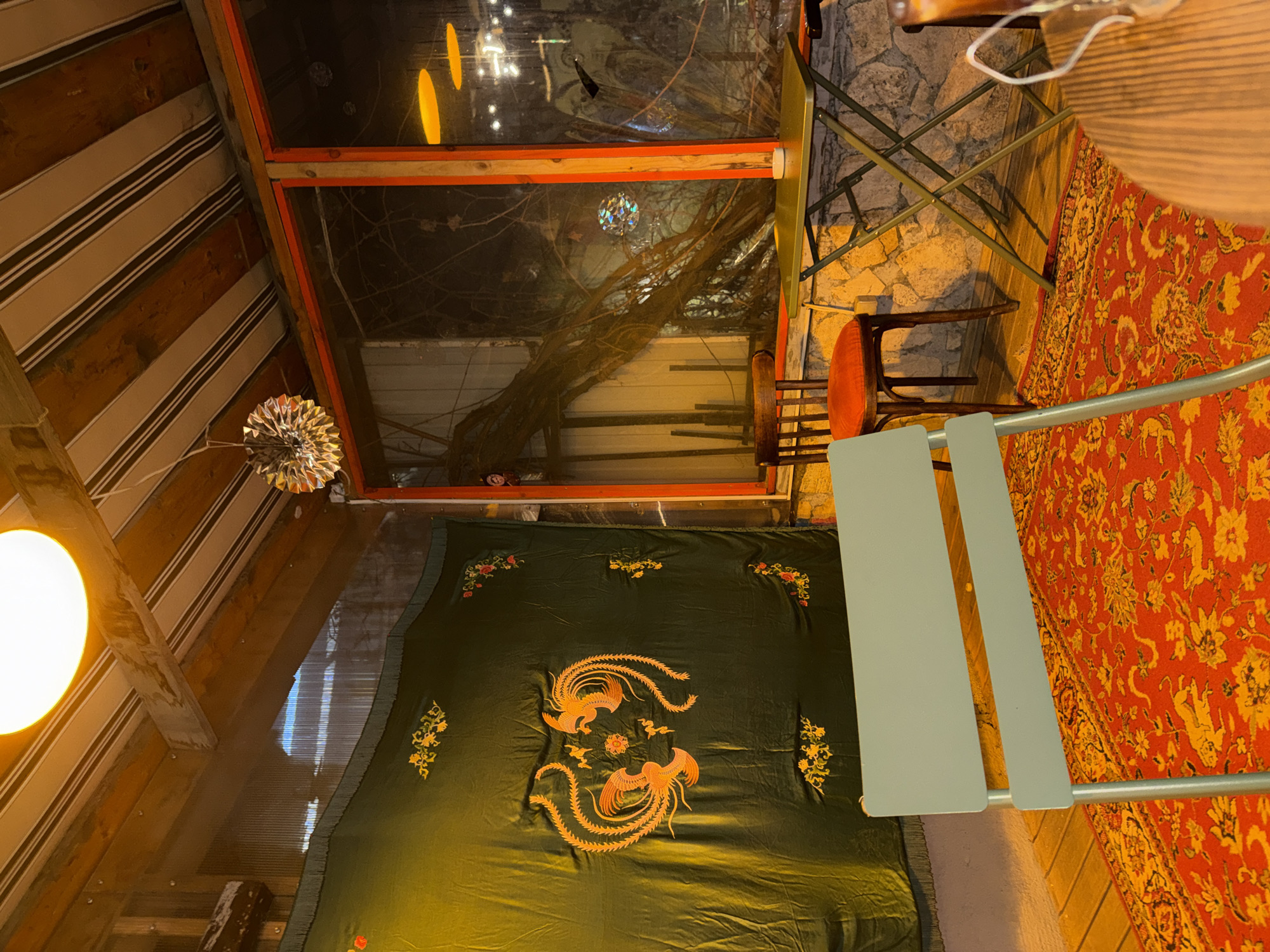 Captured indoors, the photo reveals a blend of vibrant colors and diverse objects within a room. The walls and ceiling, constructed from striped wooden panels, create a warm, textured atmosphere. The light source from the left casts an orange hue, highlighting the room’s structure.
A prominent feature is a large window with an orange frame, through which bare branches of a tree are visible, adorned with what appear to be decorative lights. Adjacent to the window is a light-green table with metal legs and an orange-cushioned wooden chair.
Below, a dark-green fabric with intricate embroidery of two golden phoenixes, complemented by floral designs, covers a portion of the floor. This is next to an orange rug covered with floral patterns. The photo captures a corner of the room, with the scene set indoors. The exact location of the photo within Yerevan, Armenia, is not discernible from the image itself.