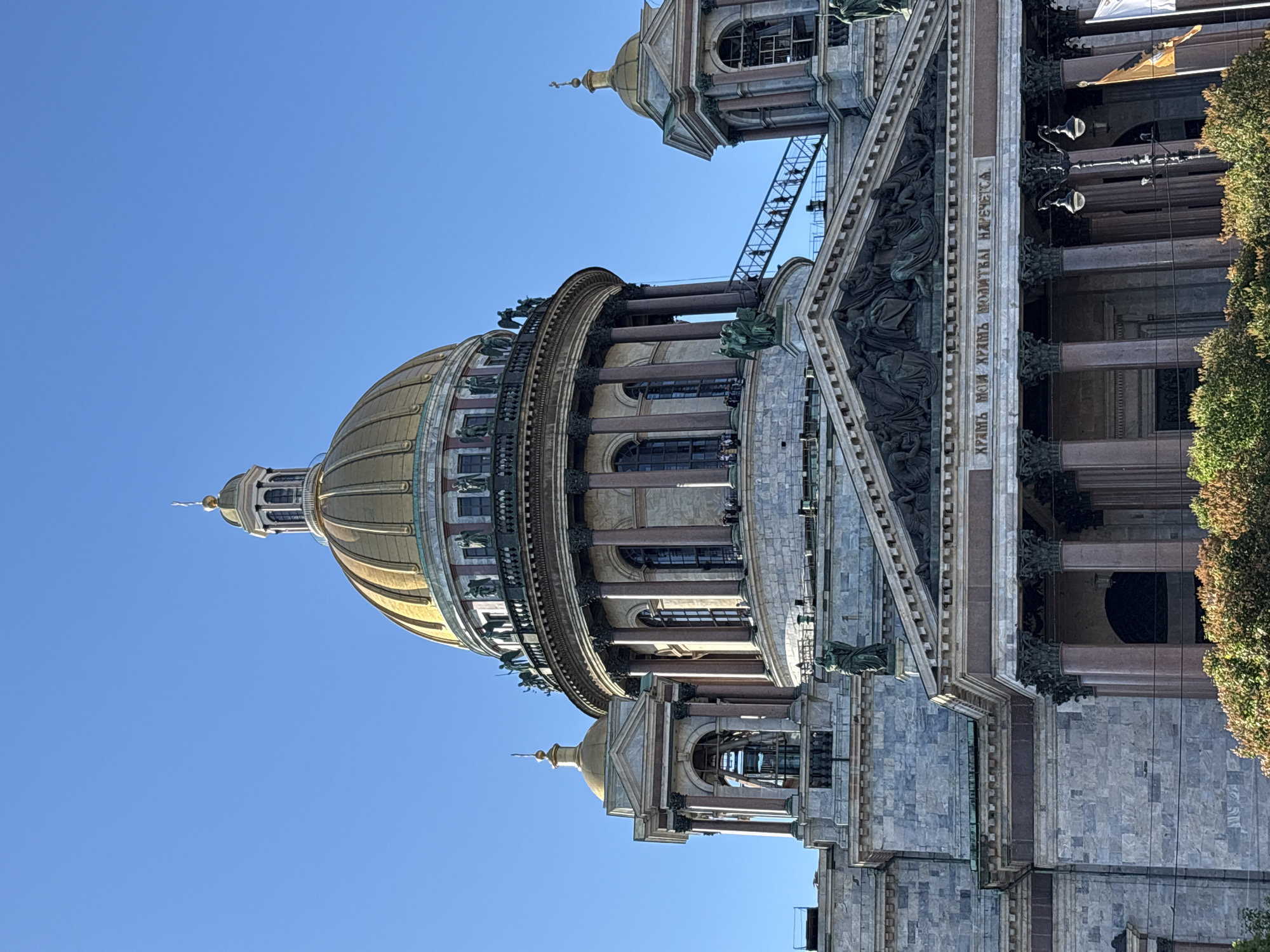 The image displays a detailed view of St. Isaac's Cathedral in Saint Petersburg, Russia, under a clear, bright blue sky. The central feature is the cathedral's massive golden dome, exhibiting distinct vertical ribbing, topped by a smaller golden cupola culminating in a spire and a cross. Below the main dome, a drum structure with numerous dark, possibly polished granite, columns and large arched windows is visible. A balustrade adorned with statues encircles the base of the dome, where several individuals are discernible on an observation deck.
To the left, one of the cathedral's smaller bell towers, also featuring a golden dome, columns, and an open belfry, rises alongside the main structure. A metal staircase, ascending towards the observation deck, is visible on the right side of the drum, with several people ascending it.
The lower visible portion of the cathedral showcases a prominent architectural facade. A large pediment, filled with intricate bas-relief sculptures depicting multiple figures, sits above the main entrance. Below the pediment, an inscription in Cyrillic reads "ХРАМЪ МОЙ ХРАМЪ МОЛИТВЫ НАРЕЧЕТСЯ" (My house shall be called a house of prayer). This section is supported by large, dark, fluted columns with ornate Corinthian capitals. The primary construction material for the cathedral's walls appears to be large, light-colored, mottled grey stone blocks. Several individual statues are positioned on various ledges and architectural elements of the facade.
In the immediate foreground, the top sections of green and brown shrubbery partially obscure the very bottom of the cathedral. A black, ornate streetlight with two lamps is visible on the right, partially behind the shrubs. To the far right, a yellow/orange flag with an emblem and a white flag are partially visible. Thin, dark power lines horizontally traverse the lower-mid section of the image in front of the cathedral. The overall lighting suggests direct sunlight.