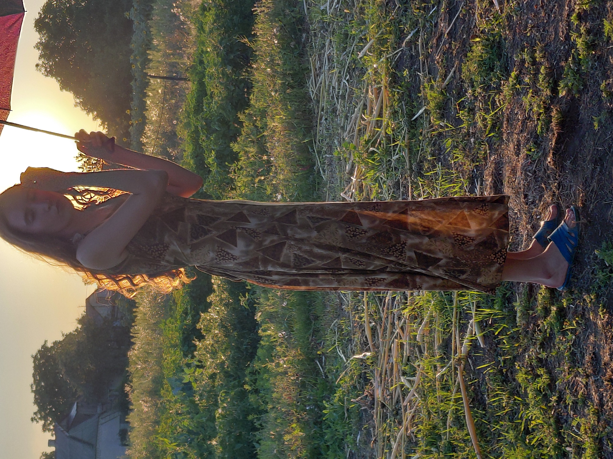 A young woman stands in a field at sunset, holding an umbrella. She is wearing a long, patterned dress and blue sandals. The field is covered in grass and dried stalks, with some vegetation in the background. The sun is setting, casting a warm glow over the scene. There are buildings visible in the distance, suggesting a rural or suburban area. The overall impression is one of peacefulness and natural beauty.