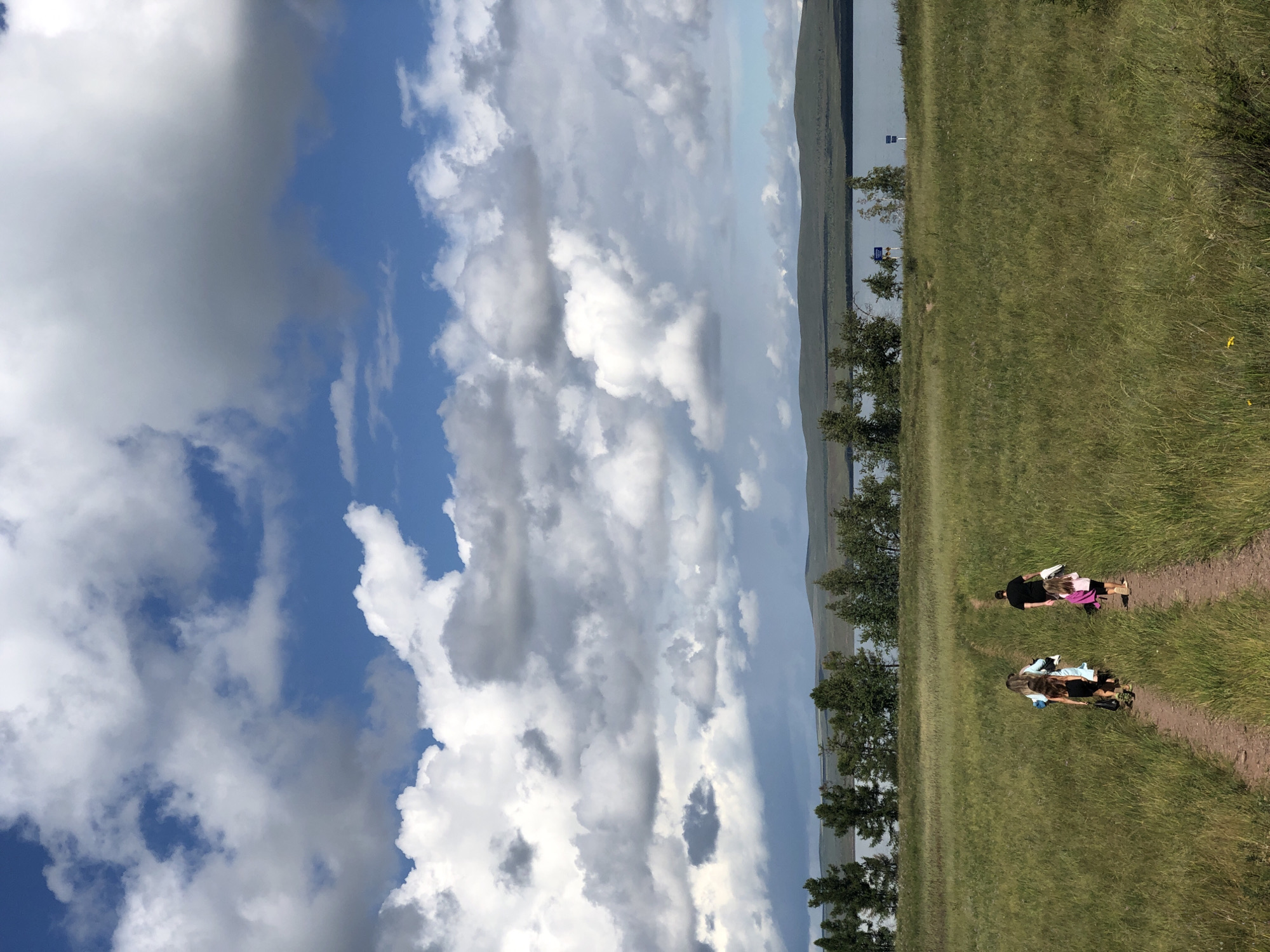 In an outdoor scene, a group of four people, two adults and two children, walk away from the camera along a dirt path through a grassy field. The adult male, in a black t-shirt, is followed by a girl in a pink shirt and shorts carrying a bag. To their left, an adult female in a light blue garment and a child in a black dress are walking ahead. The path leads towards a body of water bordered by a line of trees and distant, rolling hills under a dramatic sky filled with large, white and gray clouds. The location is the city of Zhemchuzhny, Russia.
