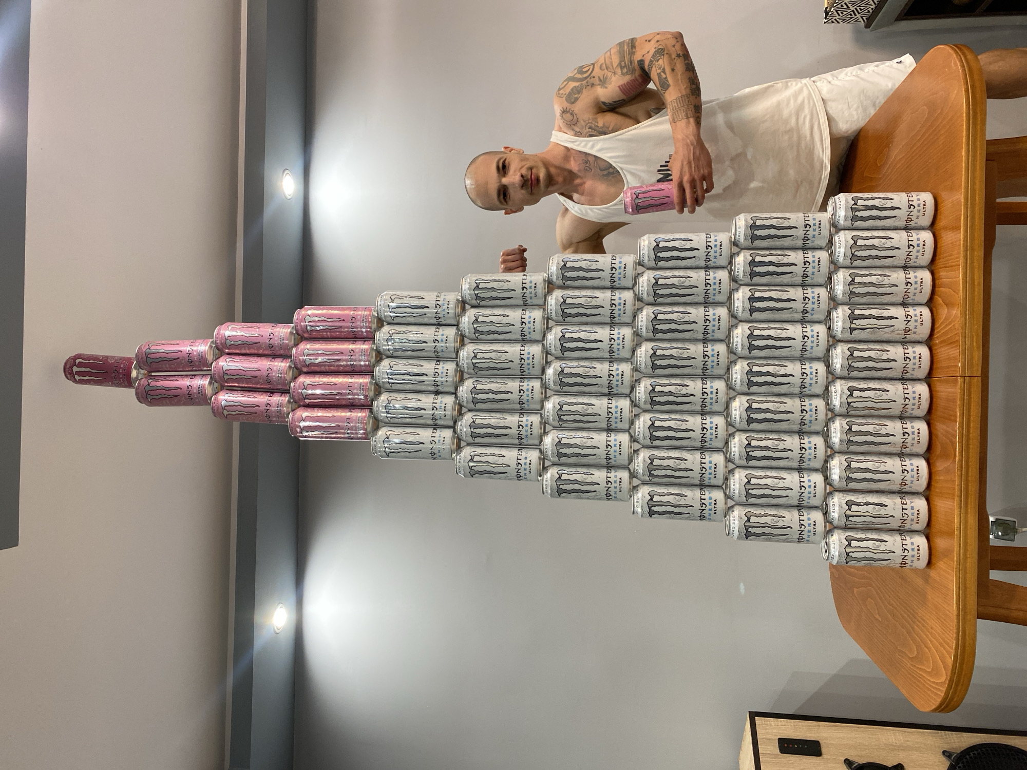 A fair-skinned, bald man with numerous tattoos on his arms stands behind a pyramid of Monster energy drink cans on a wooden table. The cans are stacked in a pyramid shape. Most are white, with a black Monster logo, while those at the peak are pink. The man is holding a pink can. He is wearing a white tank top. The background is a neutral gray wall. The location is not mentioned in the image.