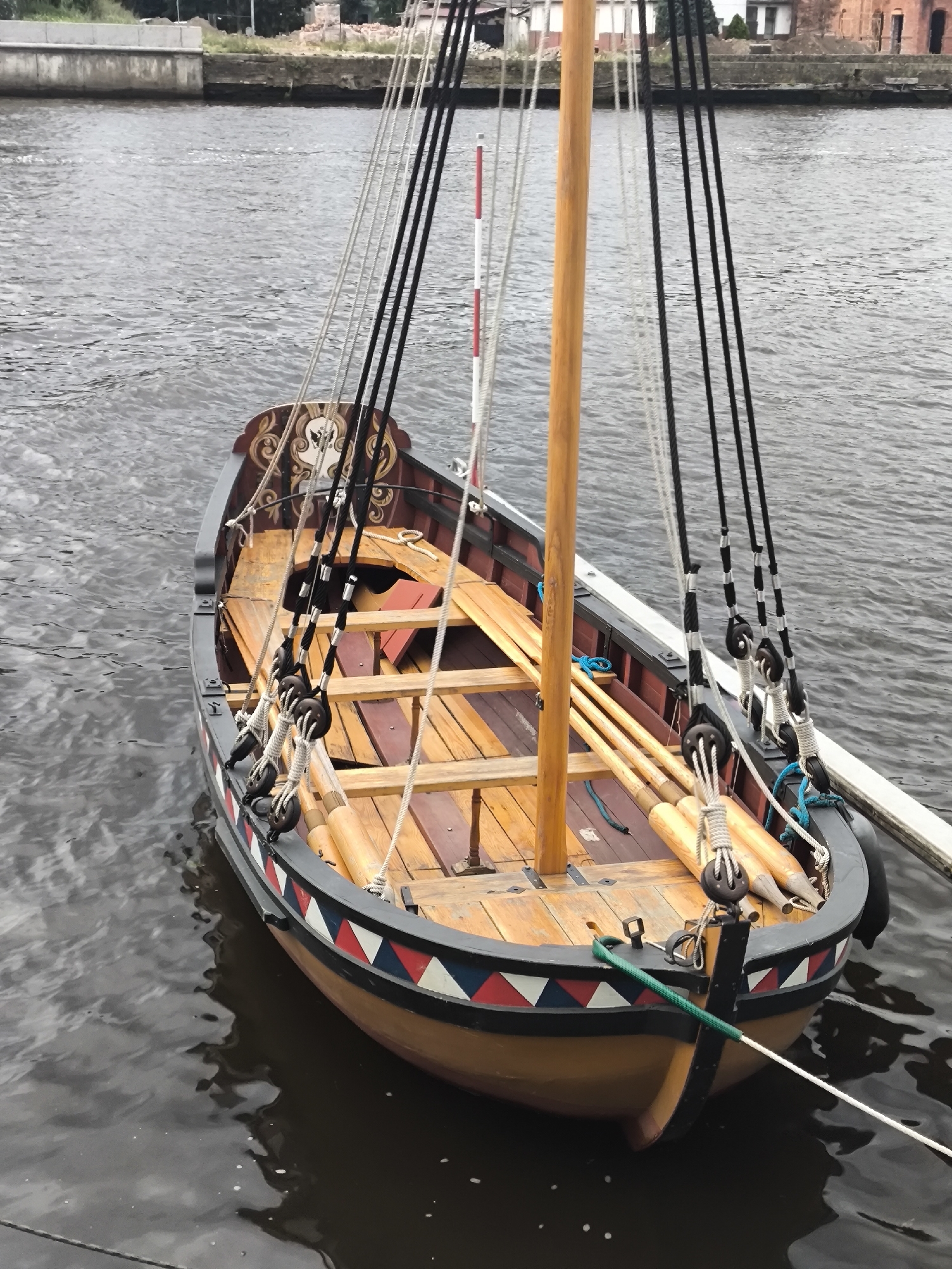 A wooden boat with a single mast is docked in dark water. The boat has a light brown hull with a decorative trim of red, white, and blue triangles. The deck is made of wooden planks, and there are several wooden benches. The boat is equipped with several ropes and pulleys, and a wooden pole extends from the boat towards the stern. Oars are stacked along the sides of the boat. In the background, a concrete embankment and some buildings are visible, suggesting an urban or industrial waterfront. This scene is likely located in Kaliningrad, Russia, possibly along the Pregolya River or in the port area.