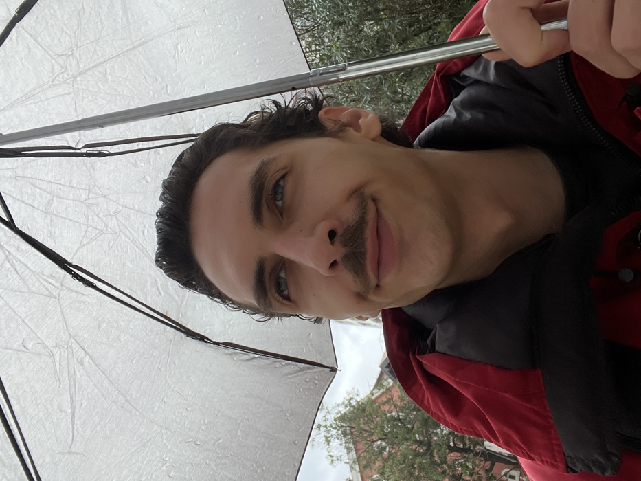 An upward-facing, chest-level portrait captures an adult male in Rafailovići, Montenegro, holding an open, clear or translucent umbrella. The individual has dark, wavy hair, blue eyes looking upward and slightly right, and a prominent mustache, displaying a slight smile. He is wearing a red jacket with a black collar. Water droplets are visible on the umbrella's surface, indicating precipitation. The background features blurred green foliage, a building with a reddish roof and light-colored walls to the left, and additional greenery to the right, under an overcast sky. The person's right hand is visible gripping the umbrella's handle in the lower right corner.
