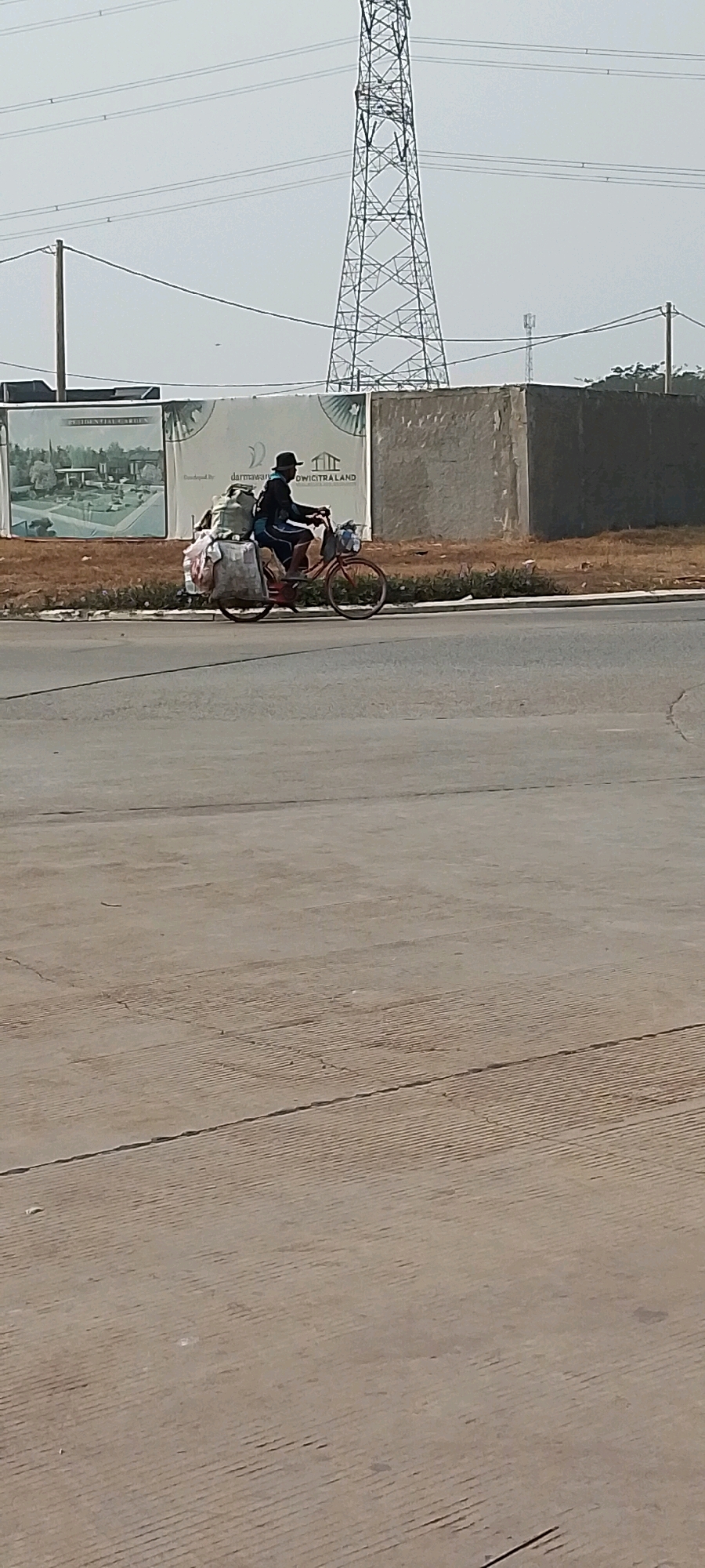 A lone individual is riding a red bicycle loaded with numerous bundles of belongings. The individual is wearing a dark shirt, light-colored pants, and a dark hat. The bicycle's rear rack is heavily laden with large, cloth-wrapped packages, and there are additional items secured to the front and possibly the sides of the bicycle. The person is in motion, pedaling the bicycle along a paved road.
The background features a large, lattice-like power pylon dominating the sky, with high-voltage power lines stretching across the frame. Behind the road, there is a low, rough-textured concrete wall and a grassy embankment. A large billboard or advertisement panel is visible behind the wall, featuring images of what appear to be residential properties and text that includes "RESIDENTIAL GARDEN" and "DERMAWAN". The overall environment suggests an industrial or developing area, possibly on the outskirts of a city. The ground in the foreground is a light-colored paved surface with visible expansion joints and a textured pattern.
The image was taken in Satriamekar, Indonesia, as indicated by the location information. The scene depicts a common sight of individuals transporting their possessions by bicycle in areas where personal transportation may be limited. The lighting suggests it is daytime, possibly late morning or early afternoon, given the brightness and the angle of any potential shadows, though clear shadows are not prominent.