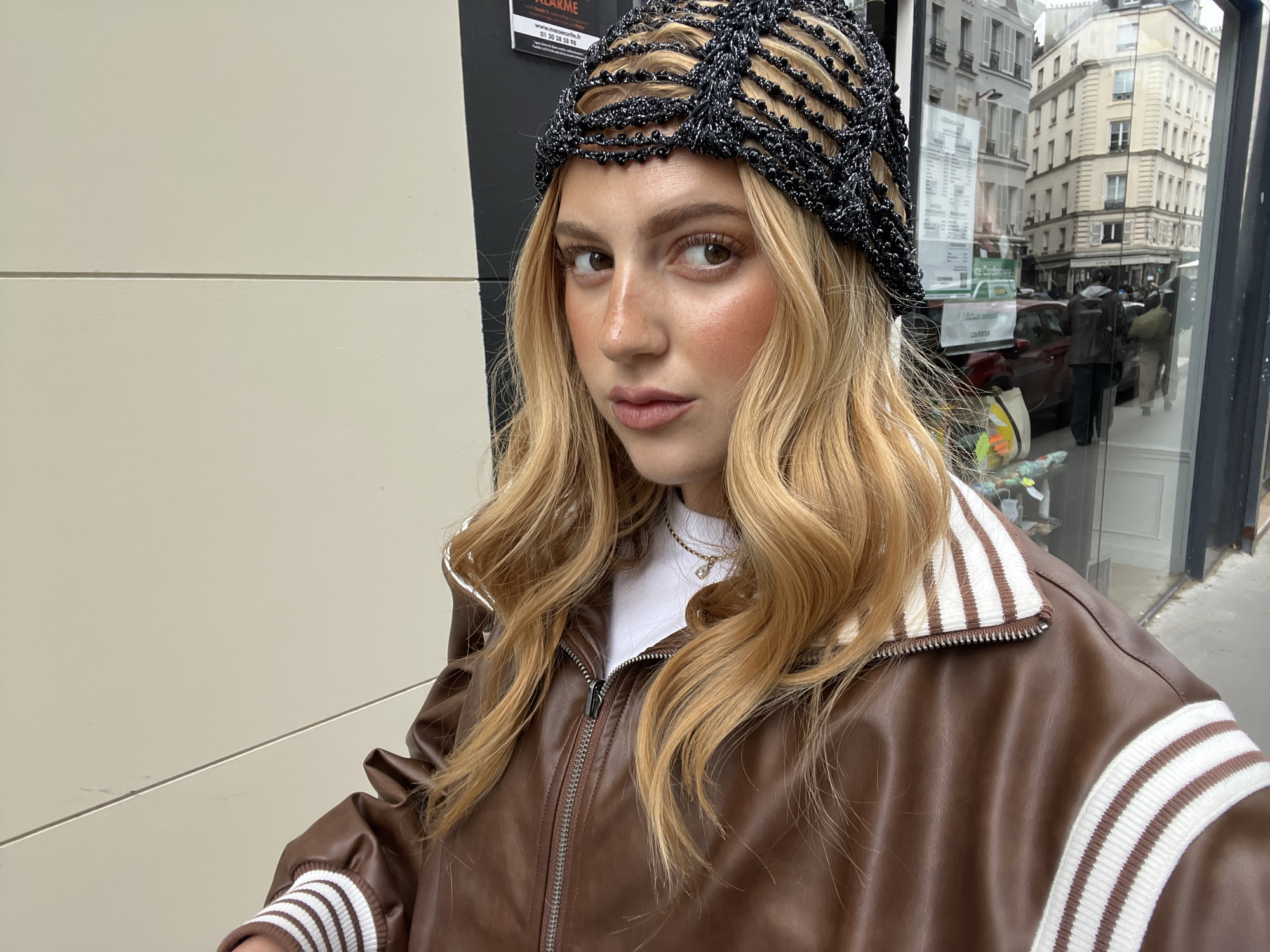 A young woman with light brown hair and brown eyes stands in front of a building in Paris, France. She wears a black netted hat, a white shirt, a gold necklace, and a brown leather jacket with white striped details on the sleeves and collar. The building behind her has a gray exterior with white panels. Reflections in a nearby window show Parisian architecture, including buildings with multiple stories and windows. A red car and several pedestrians are visible in the reflection.