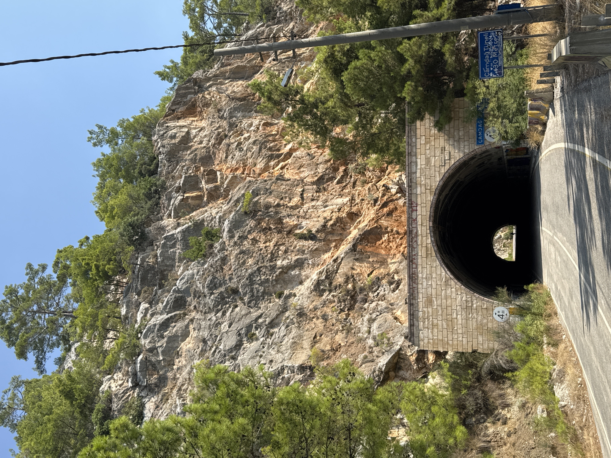 The image shows the entrance to the Çamdağ Tunnel in Kemer, Turkey, carved into a rocky mountainside. The tunnel is constructed with a stone facade and an arched opening. A paved road leads up to the tunnel, with white lines marking the lanes. On the left side of the road, a circular sign indicates a height restriction of "4.00". On the right side of the road, a blue sign with white lettering reads "Çamdağ Tüneli" and "30 Metre". A utility pole with a lamp is situated to the right of the tunnel entrance. The mountainside is covered with trees, primarily pine trees, and the sky is clear and blue. The overall scene depicts a mountain road infrastructure in a natural, mountainous environment.